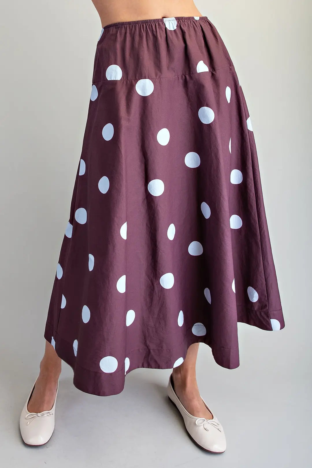 A person wearing a high-waisted, A-line midi skirt in a deep plum purple with a white polka dot pattern, paired with white ballet flats, against a plain light gray background.