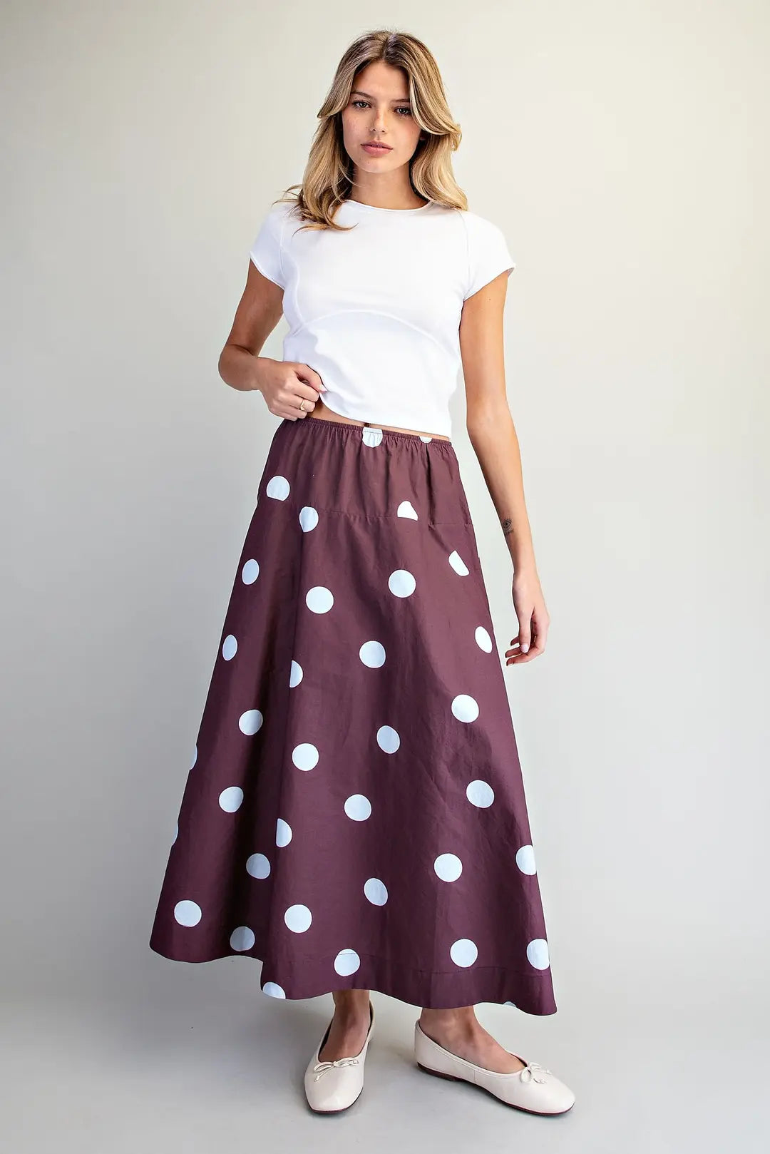 A woman modeling a long, flared skirt with large white polka dots on a deep plum-purple background, paired with a white t-shirt and white flats, against a neutral light gray studio background.