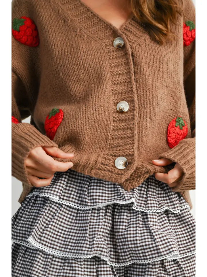 A close-up of a person wearing a brown ribbed knit cardigan with crocheted strawberry patches and silver buttons, paired with a black-and-white gingham ruffled skirt.