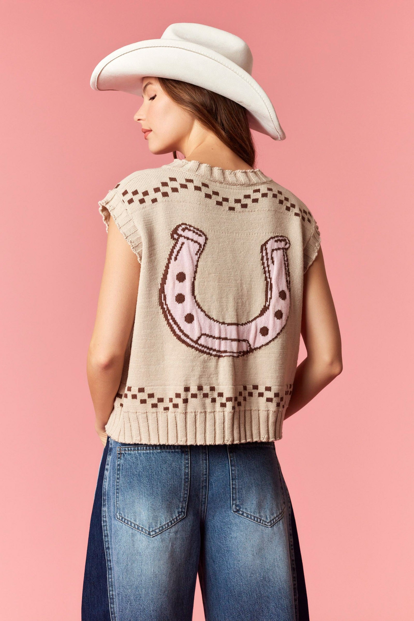 Beige knitted vest with a large pink horseshoe graphic on the back, featuring brown dotted accents and a ribbed hem, styled with blue jeans