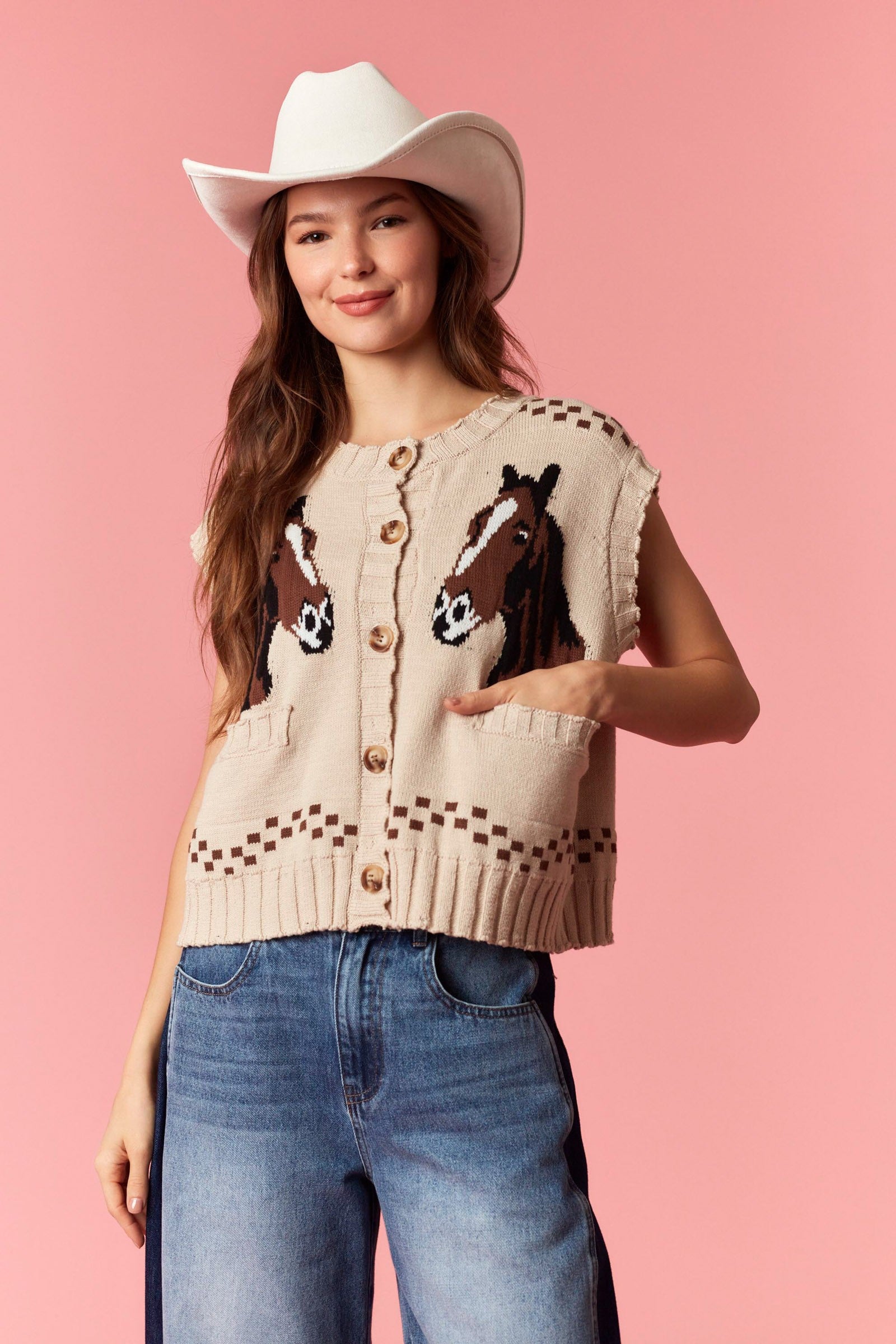 Cream knit vest with horse head and saddle motifs, button-front closure, and checkered pattern at hem, styled with blue jeans against pink background