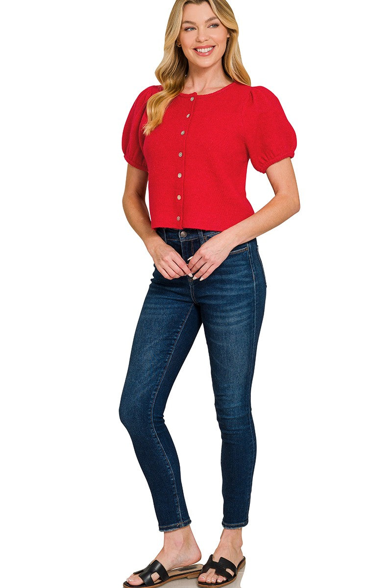 A woman modeling a bright red short-sleeve puff-sleeve top with gold buttons, paired with dark blue skinny jeans and black slide sandals, against a plain white background.