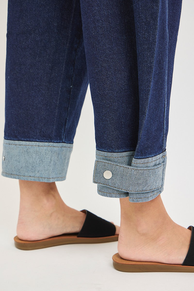 Close-up of dark blue cropped barrel cuff jeans with light wash interior, rolled at the hem with snap button detail, worn with black slide sandals