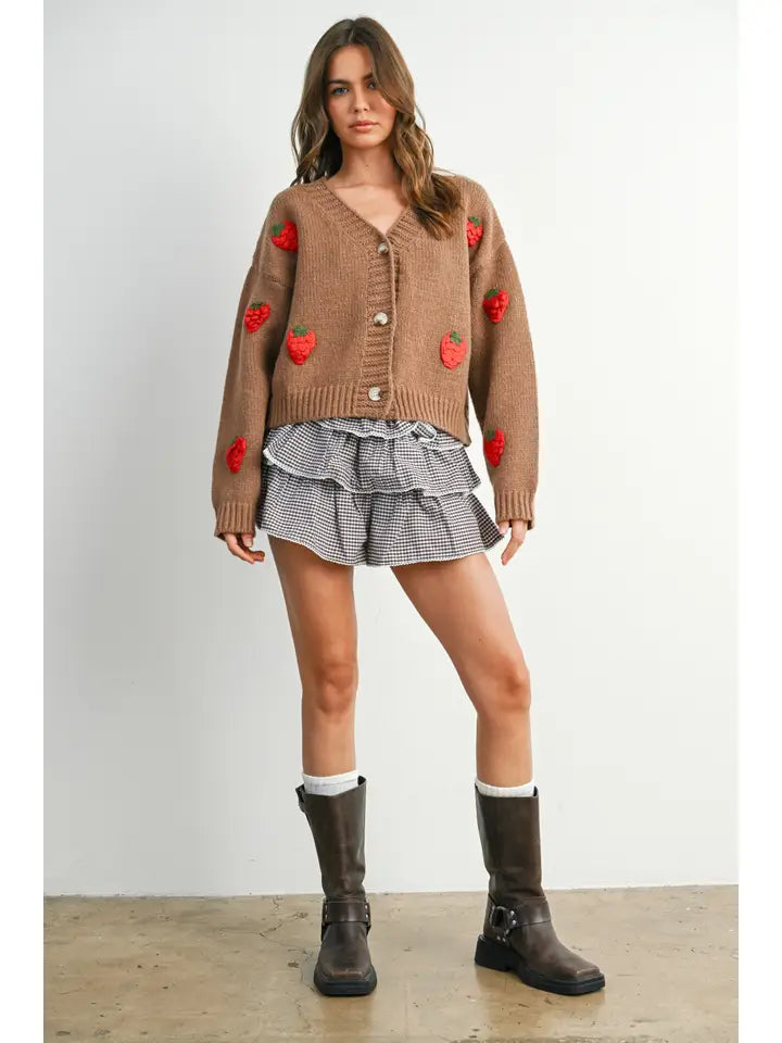 A woman modeling a brown knit cardigan with red strawberry patches, paired with a gray and white ruffled mini skirt, white socks, and dark brown boots, set against a plain light gray wall and concrete floor.