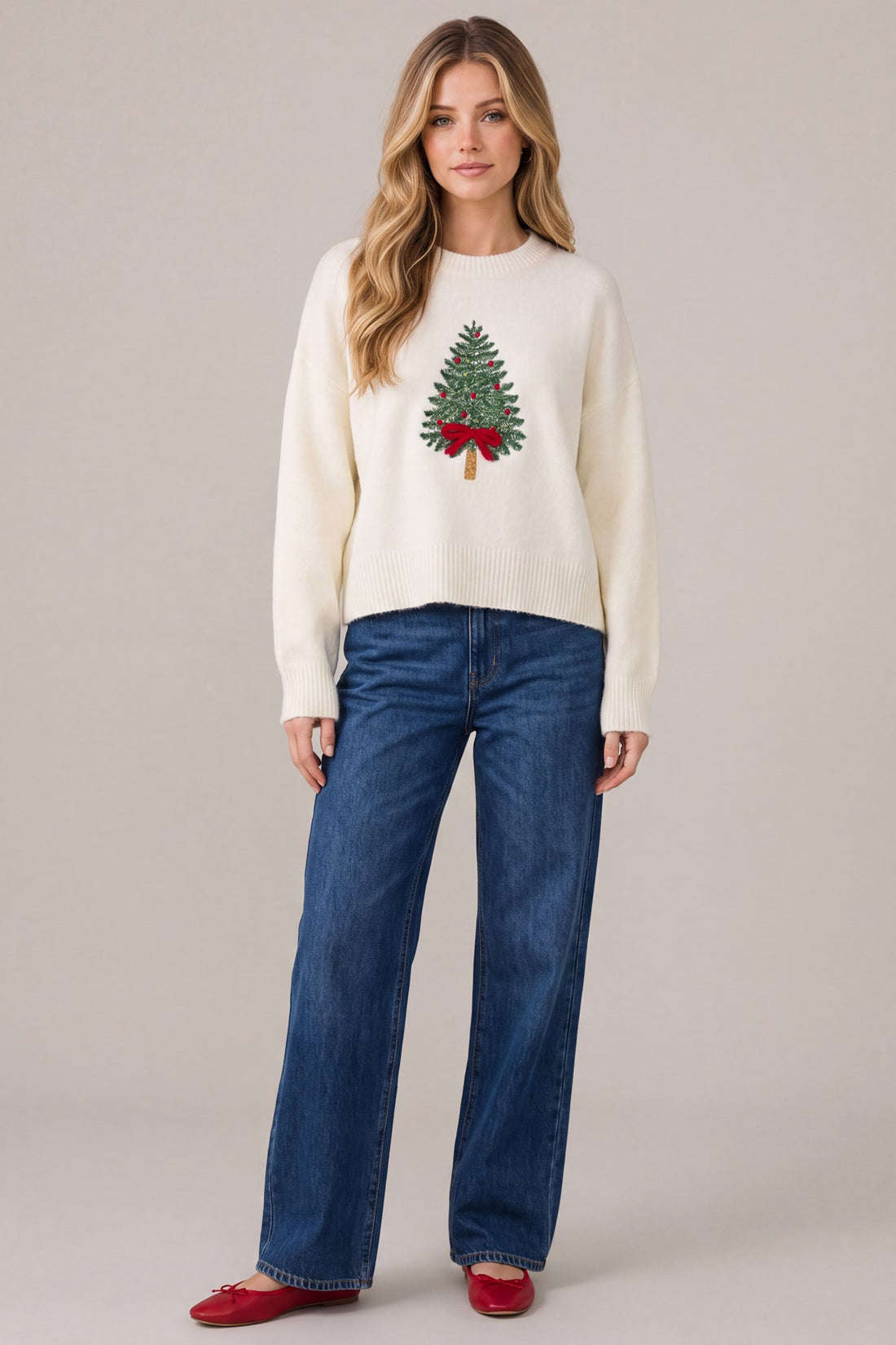 Woman sitting near a decorated Christmas tree wearing a cozy cream Christmas sweater with embroidered Christmas tree design, surrounded by wrapped gifts