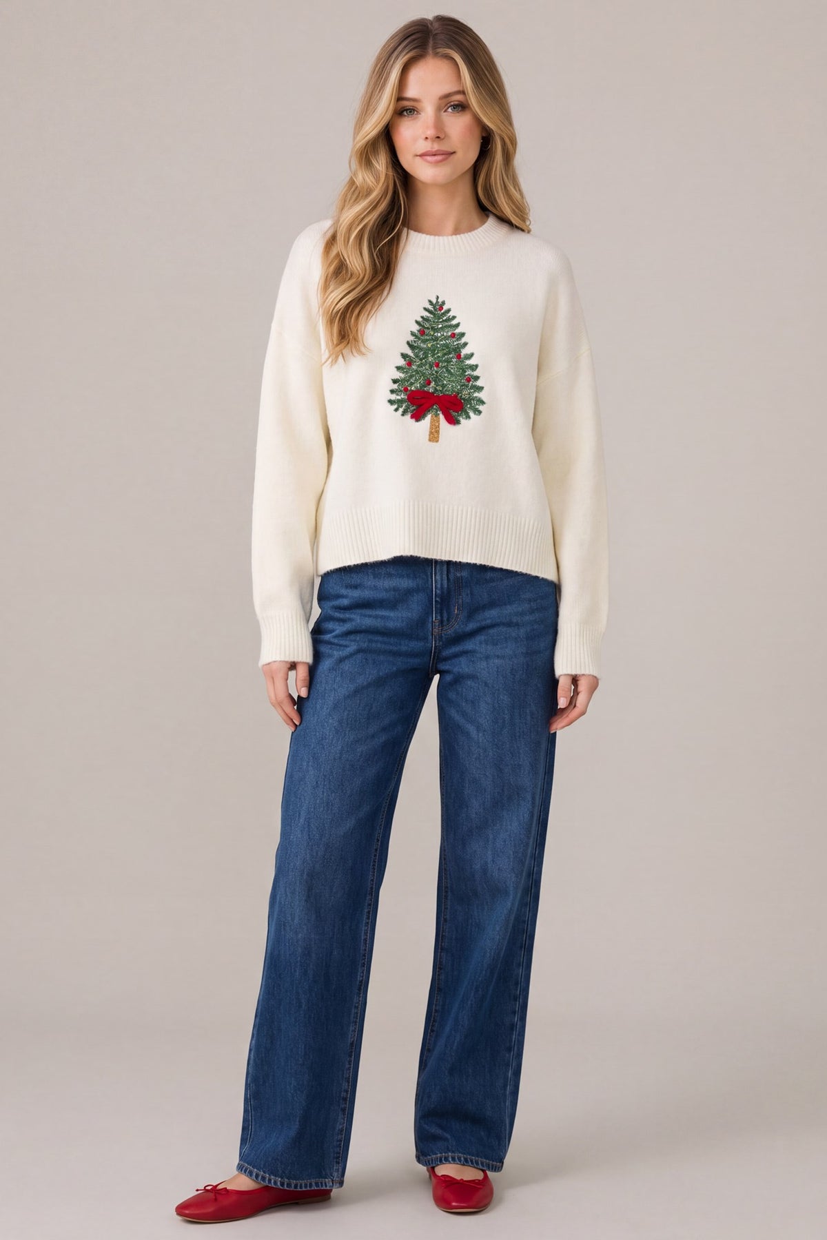 Woman standing against a plain background wearing a white Christmas sweater with embroidered Christmas tree and red bow, paired with blue jeans