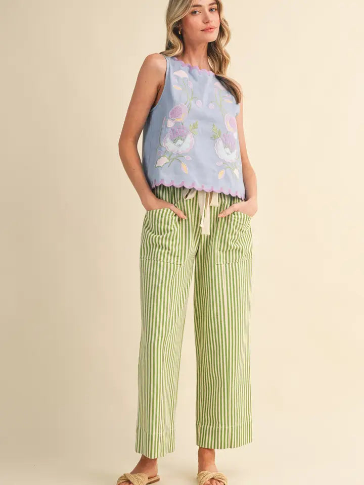 A woman modeling a sleeveless light blue top with a pastel floral print and ruffled hem, paired with light green and white vertically striped cropped pants, against a plain beige studio background.