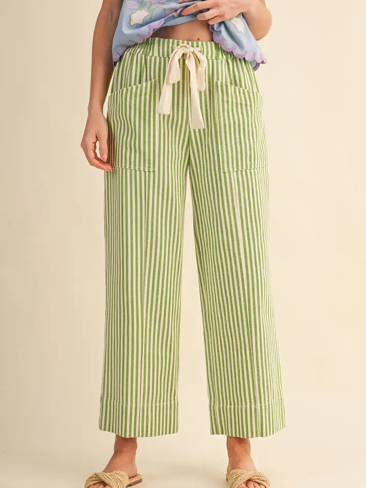 A person wearing light green and white vertically striped cropped pants with a cream drawstring waist, paired with a blue patterned top and beige slide sandals, against a plain beige studio background.