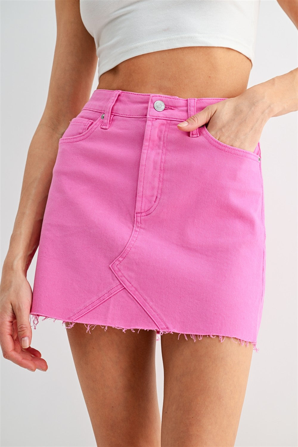 Pink high-rise mini skirt with raw frayed hem, cotton blend fabric, front pockets, and button zipper closure, styled with a white crop top.