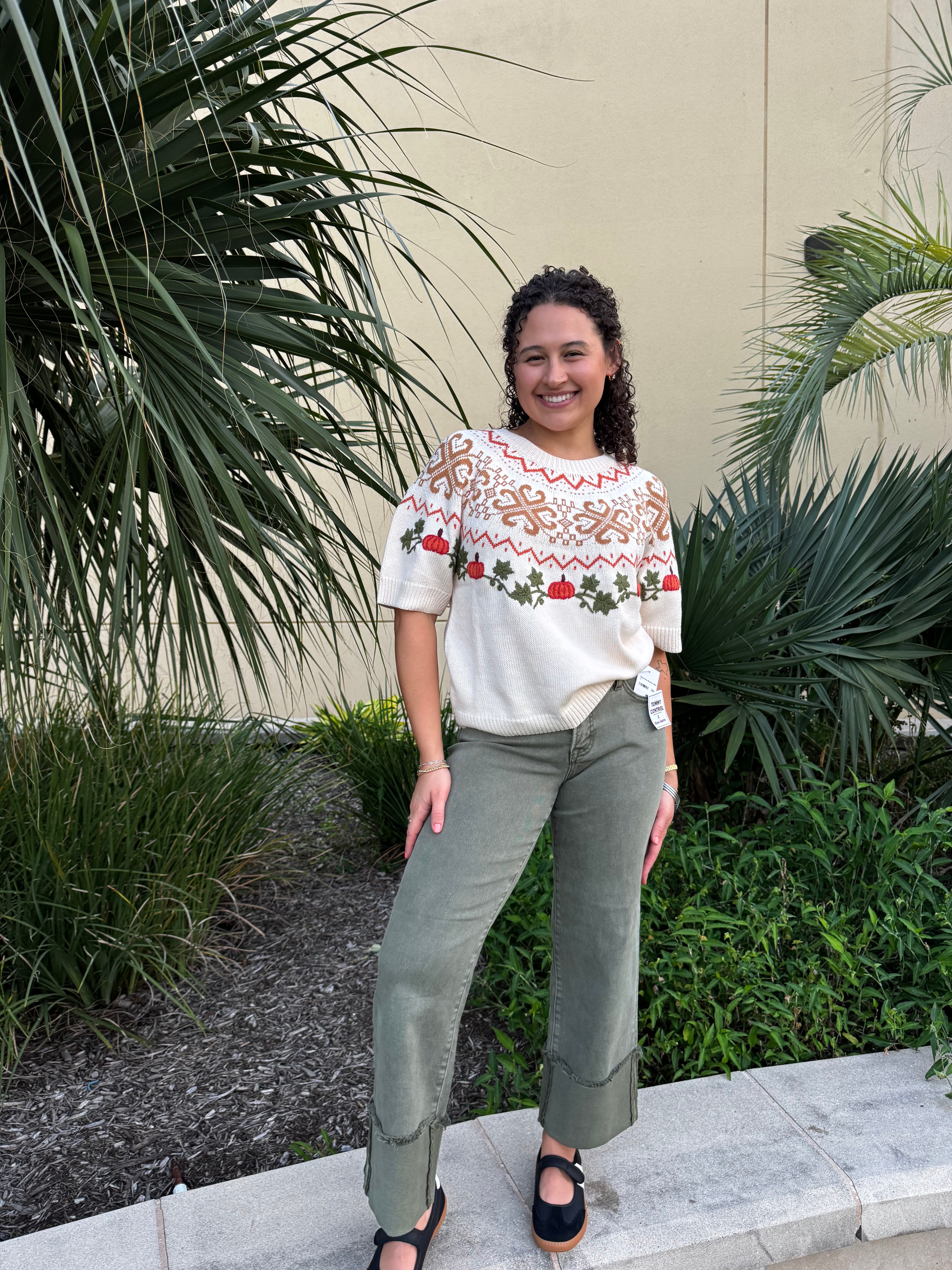 Close-up of woman with curly dark hair smiling, wearing a holiday-themed sweater with pumpkins and holly, posing outdoors near plants.
