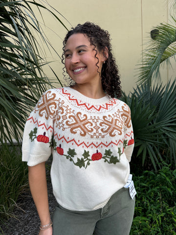 Smiling woman with curly dark hair wearing a festive cream sweater with embroidered pumpkins, holly, and intricate patterns, standing among green plants.