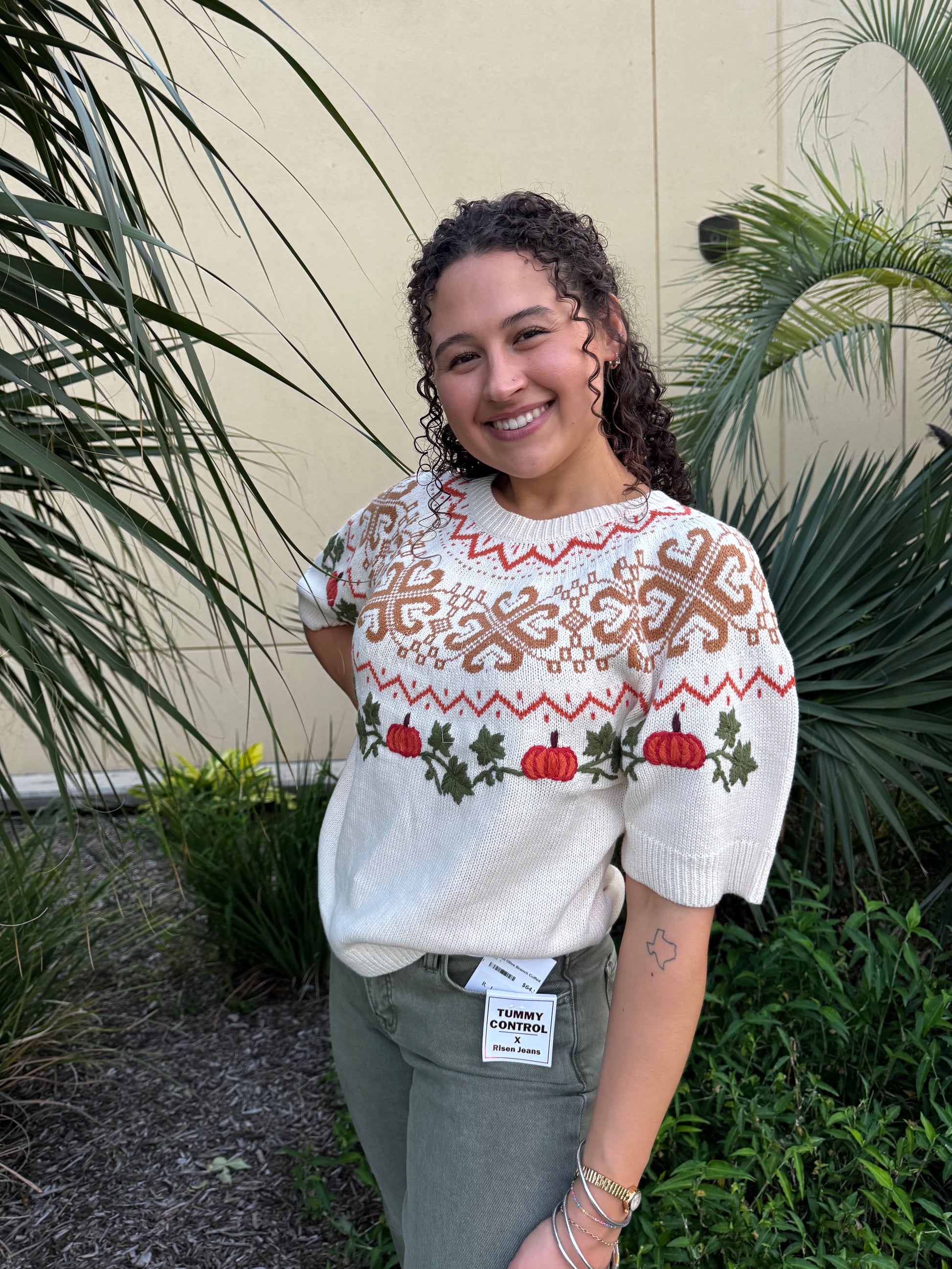 Woman with curly dark hair smiling in a festive sweater featuring embroidered pumpkins, holly, and traditional patterns, standing outdoors.