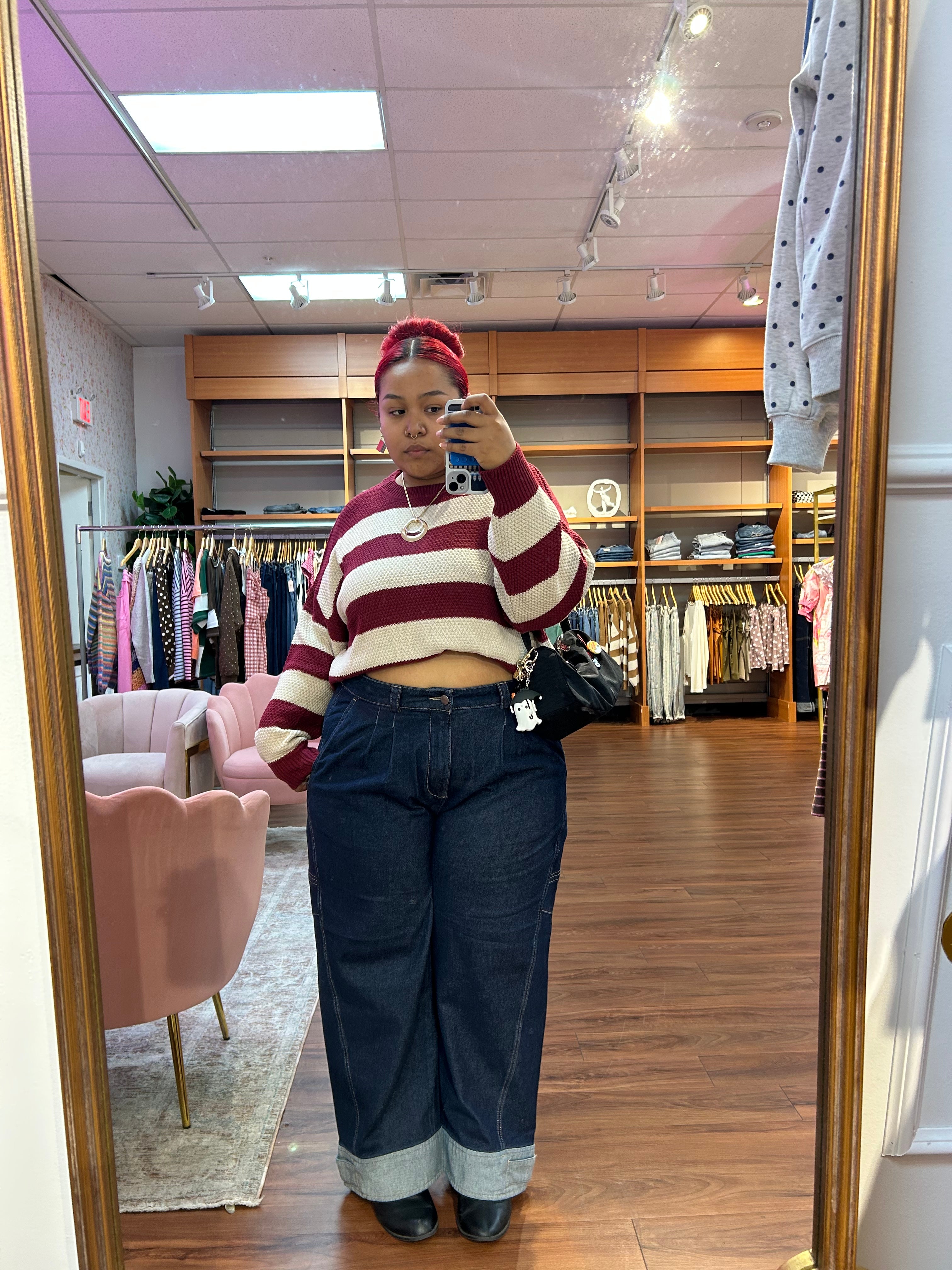A woman takes a mirror selfie in a boutique clothing store, wearing a burgundy and cream striped cropped long-sleeve top, dark blue wide-leg jeans with cuffed hems, and black shoes.