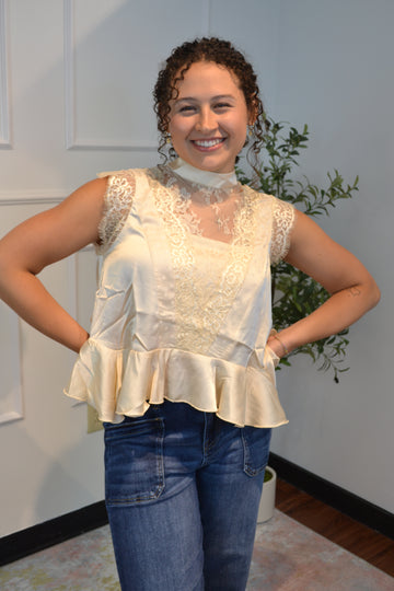 Elegant ivory satin peplum blouse featuring floral lace, sheer mesh detailing, flutter cap sleeves, and a tie-back mock neckline. Perfect for weddings, brunch, and date nights.
