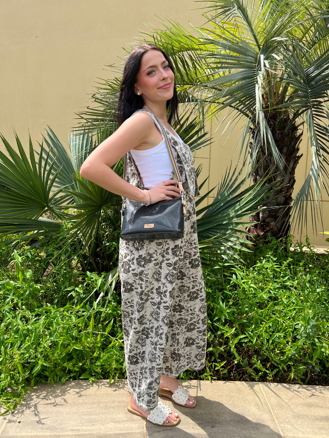 Young woman wearing floral print boho overalls with wide-leg design, white tank top, and white slide sandals, standing outdoors with lush green plants in the background.