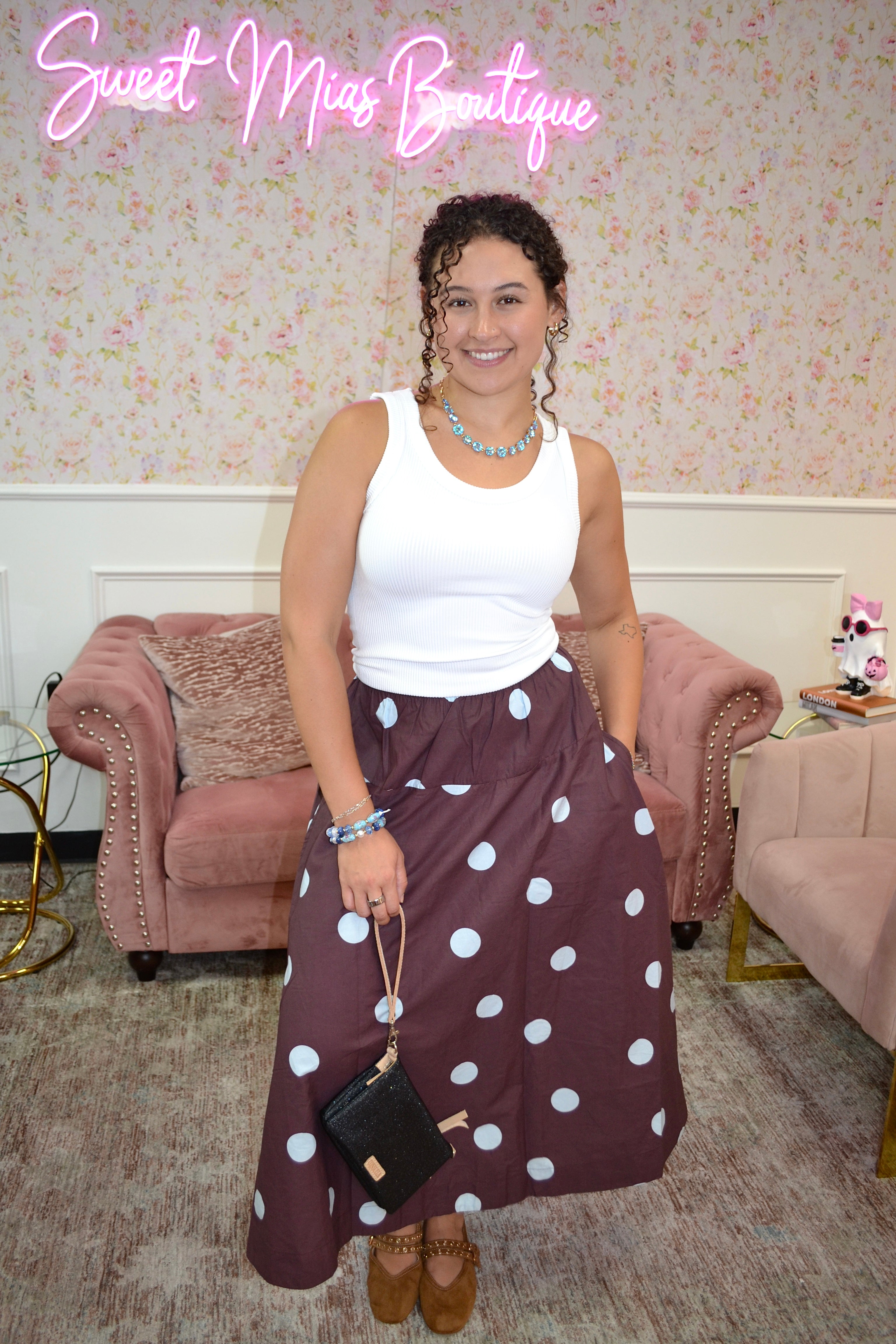 A woman posing in a white sleeveless top and a dark brown skirt with white polka dots, holding a small black handbag. The setting is a boutique interior with pink armchairs and a floral wallpaper background, featuring a pink neon sign that reads 'Sweet Mia Boutique'.