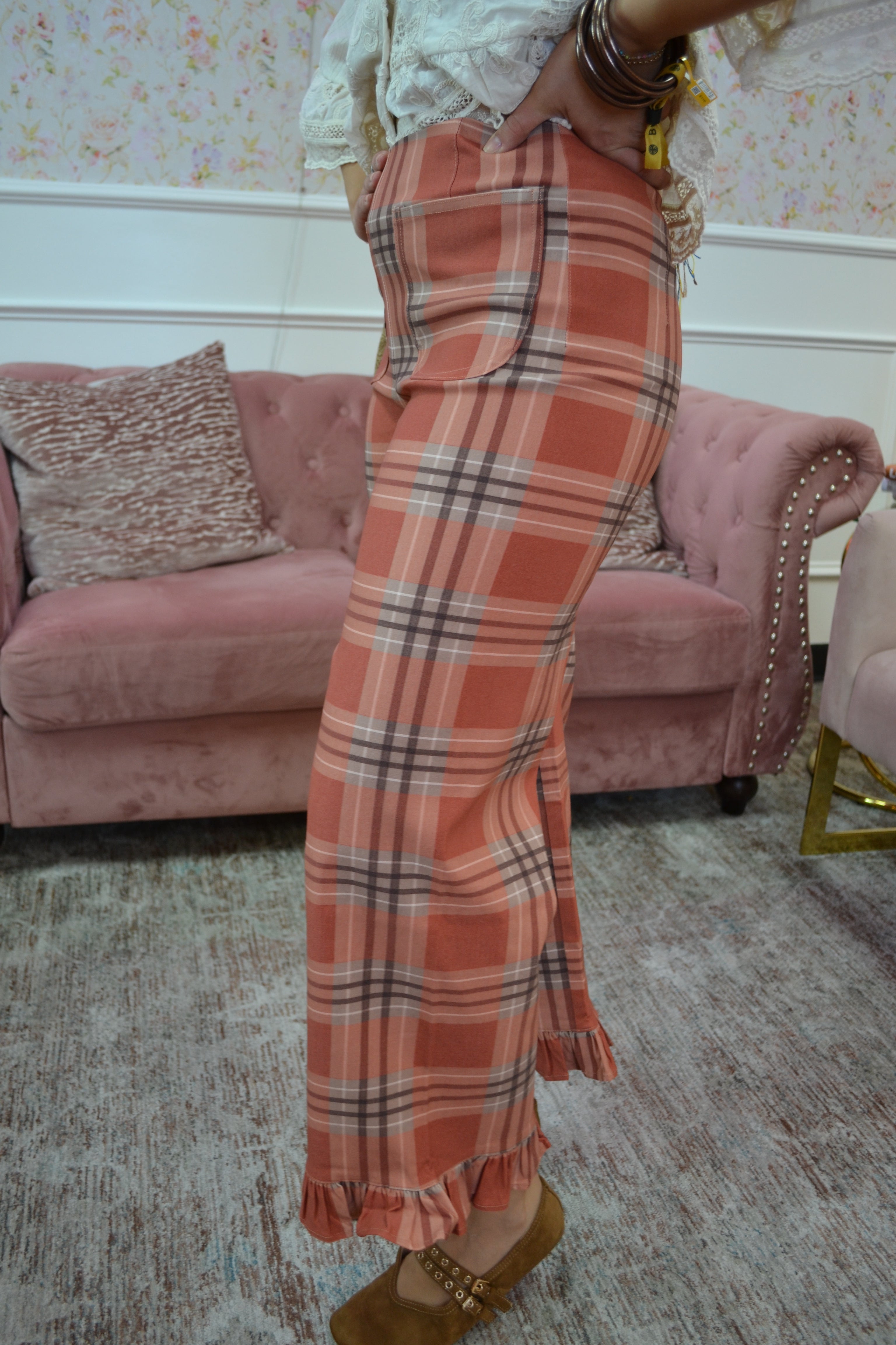 A person wearing a long, wide-leg plaid maxi skirt in a dusty orange, pink, and white tartan pattern, standing in a living room with a pink velvet sofa in the background.