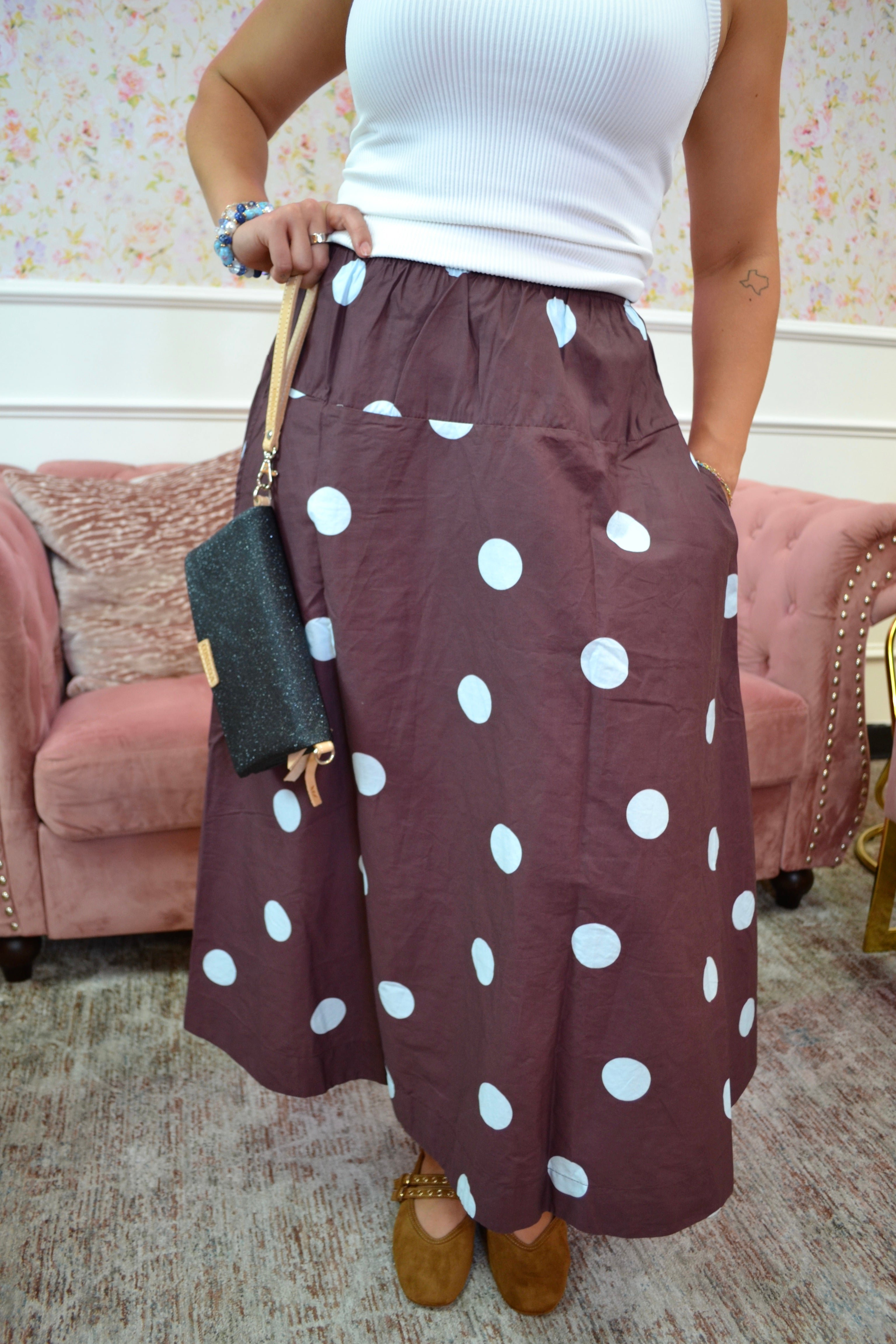 A person wearing a brown skirt with large blue polka dots, styled with a white ribbed tank top, brown suede shoes, and holding a black clutch bag