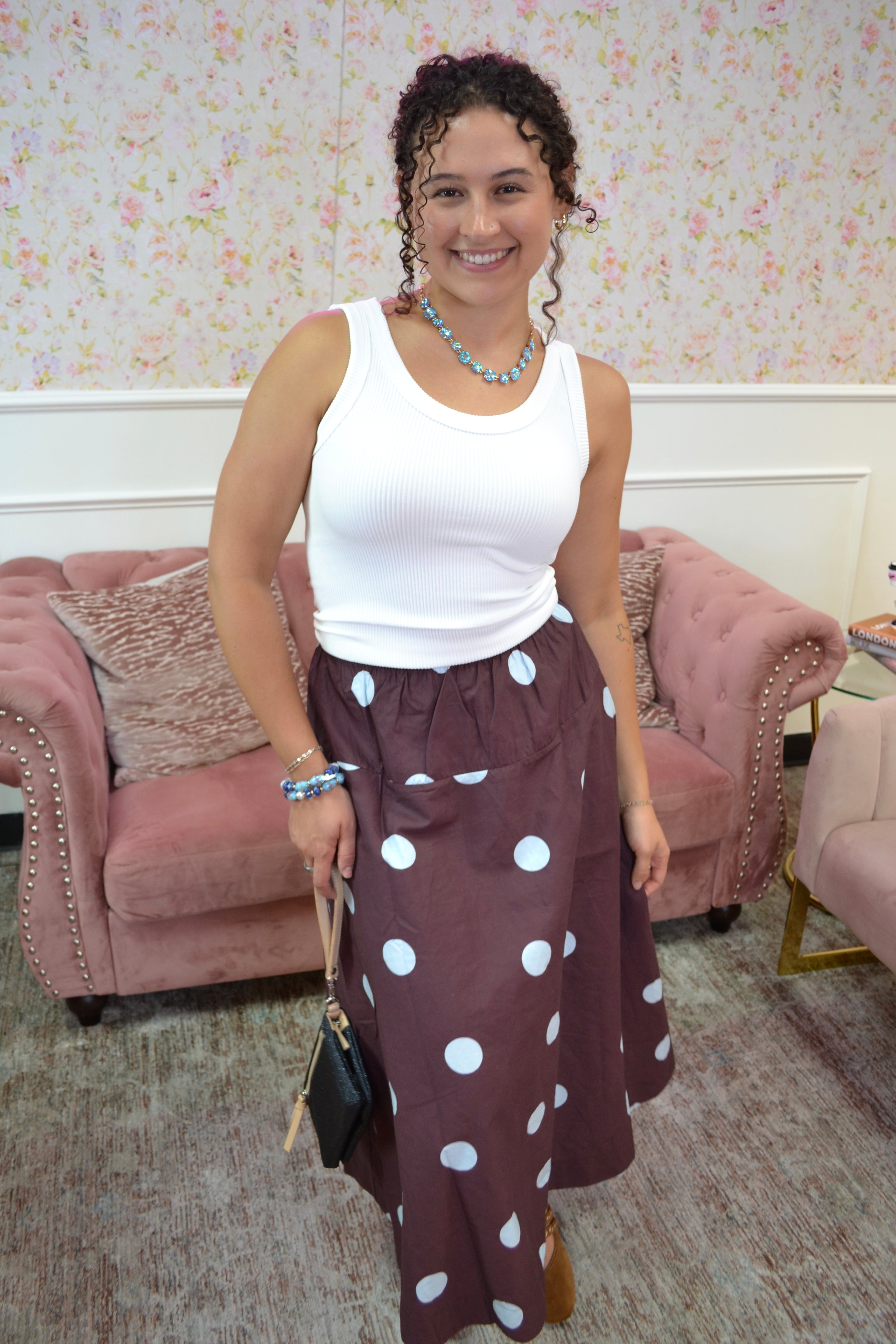 A woman posing in a white ribbed tank top and a brown midi skirt with blue polka dots, standing in a room with pink tufted sofas and floral wallpaper.