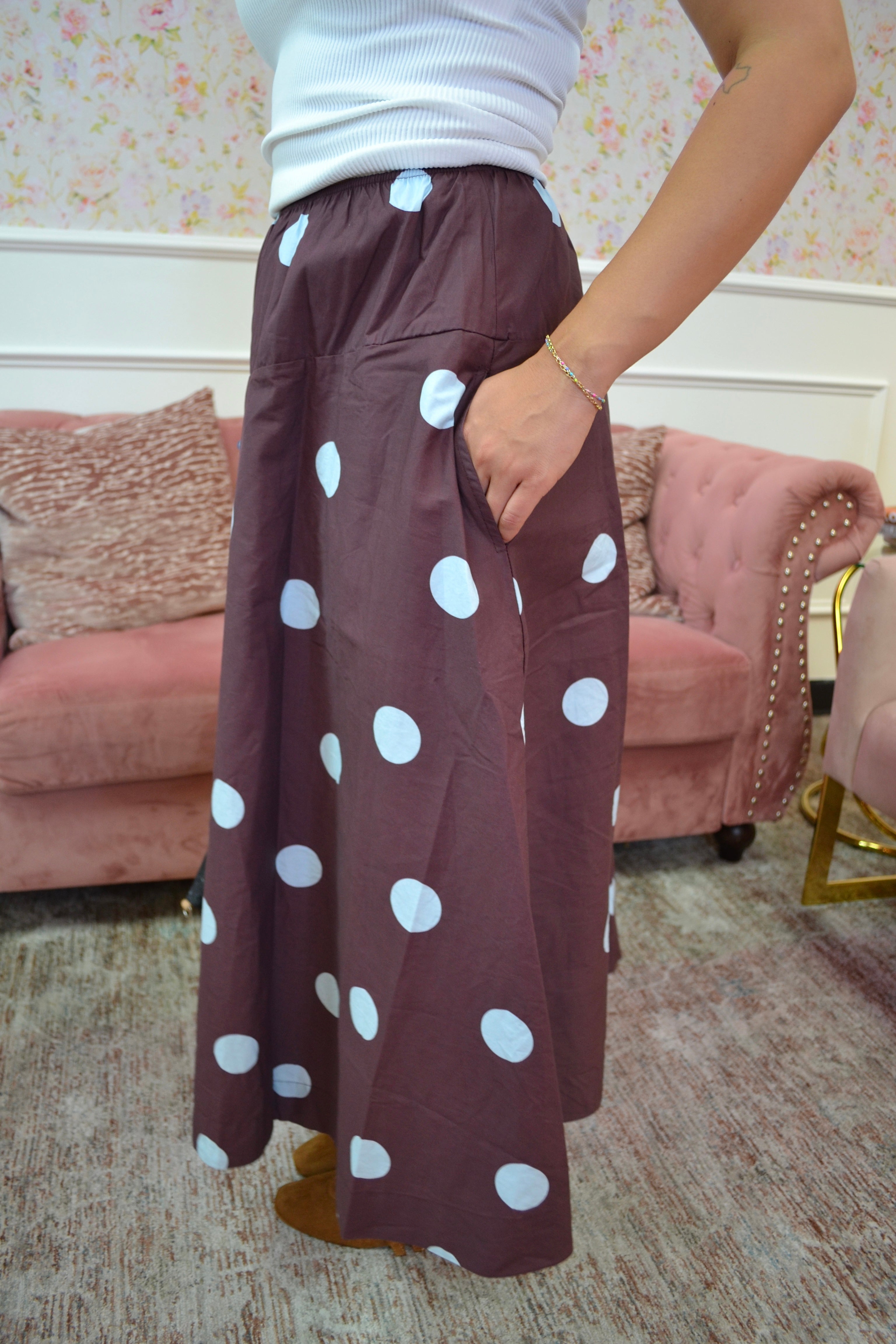 A person wearing a brown skirt with large blue polka dots, standing in a living room with a pink sofa and patterned wallpaper in the background.