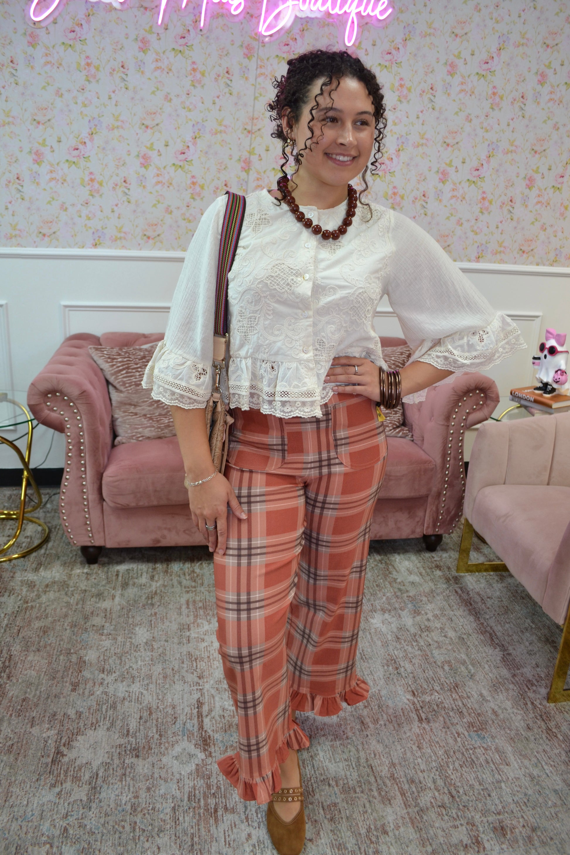 A woman posing in a boutique setting wearing a white lace-trimmed blouse and orange plaid ruffled trousers, accessorized with a brown shoulder bag and a beaded necklace.