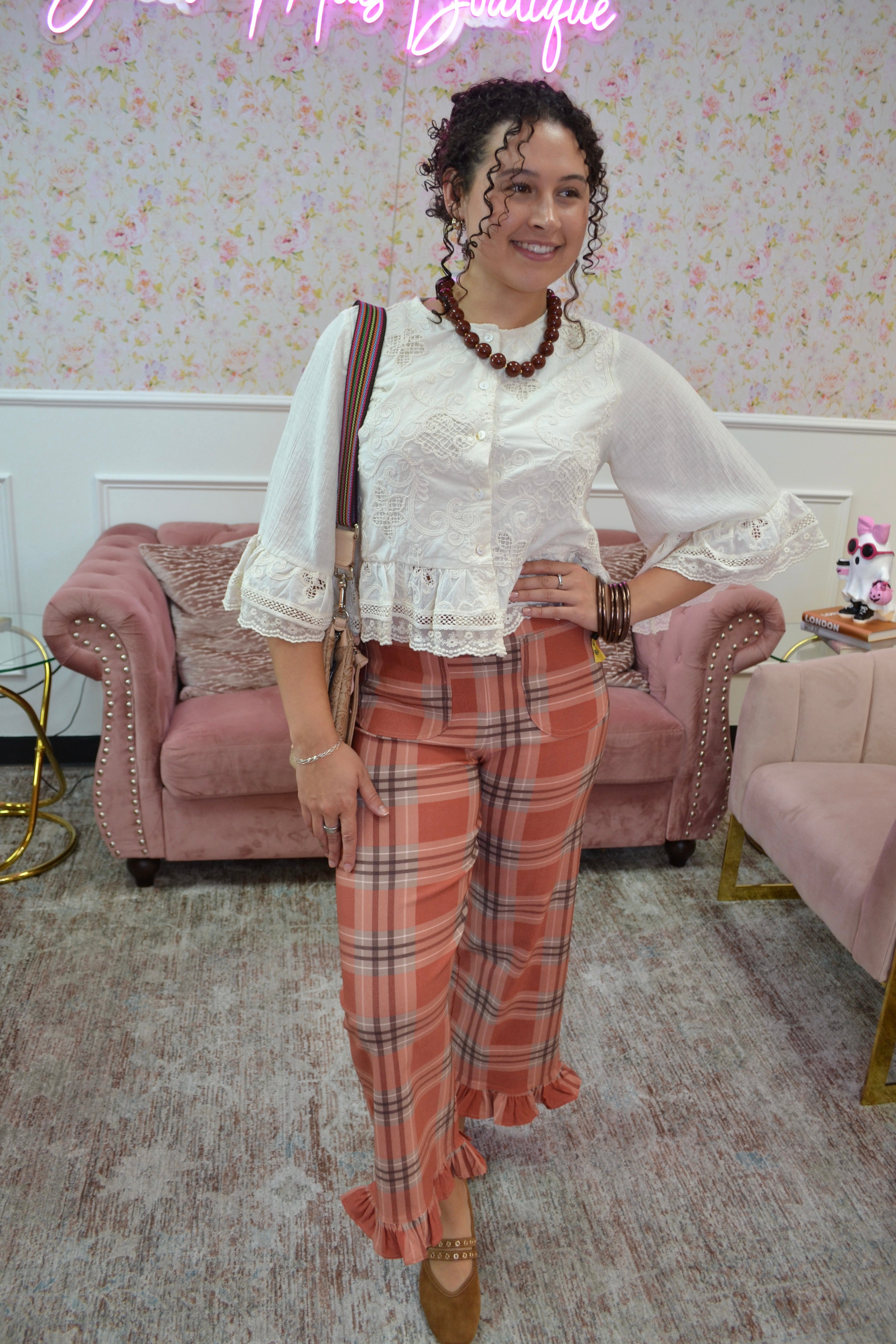 A woman posing in a boutique setting wearing a white lace-trimmed blouse and orange plaid ruffled trousers, accessorized with a brown shoulder bag and a beaded necklace.