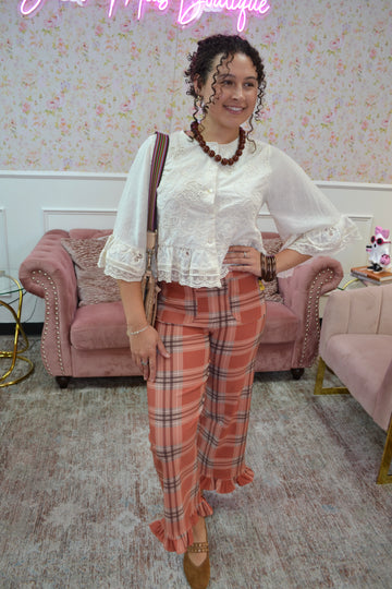 A woman posing in a boutique setting wearing a white lace-trimmed blouse and orange plaid ruffled trousers, accessorized with a brown shoulder bag and a beaded necklace.