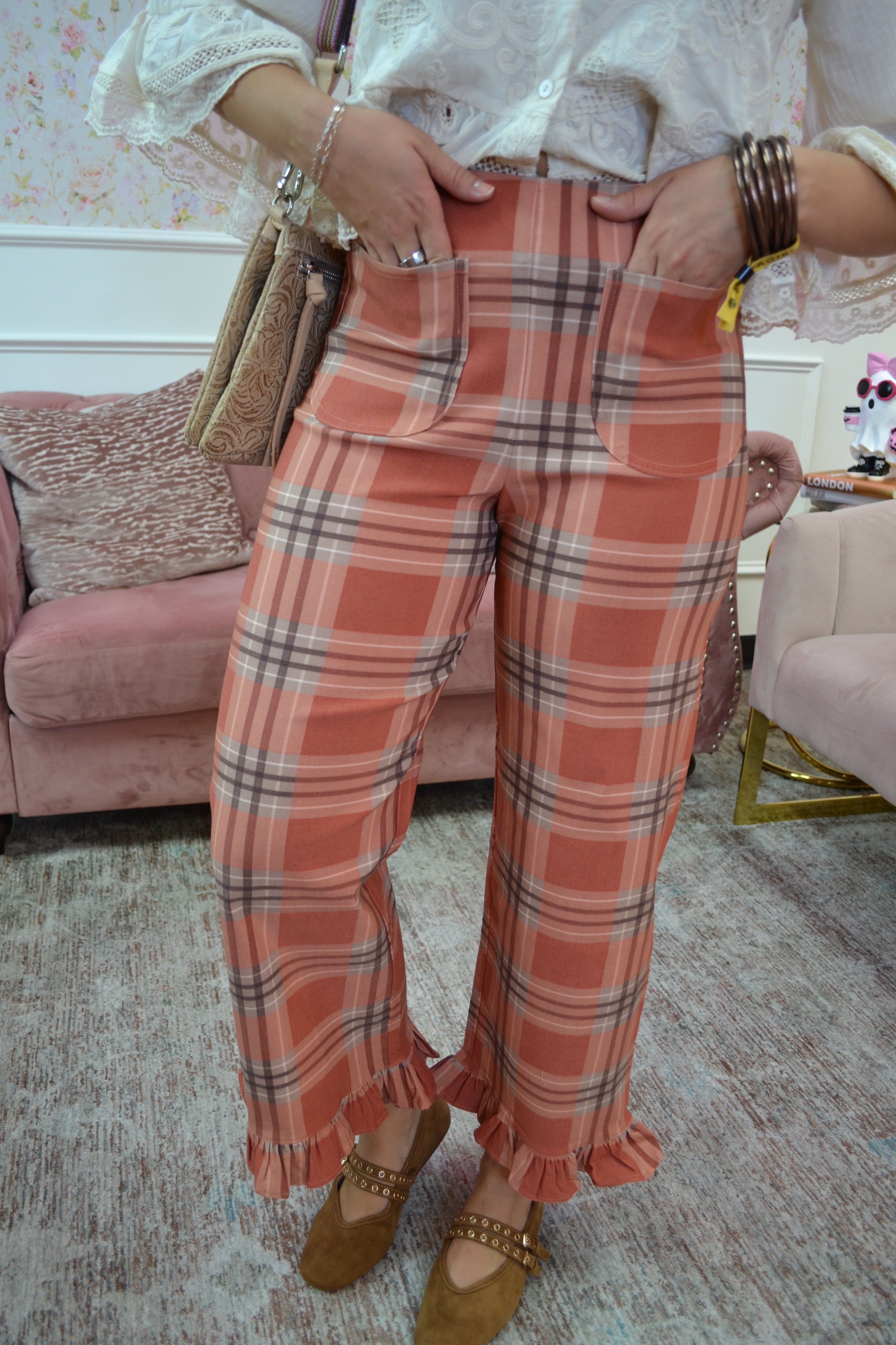 A person wearing high-waisted plaid cropped pants in a warm peach-orange tone with thin gray and white lines, paired with a white blouse and brown flats, standing in a living room setting.