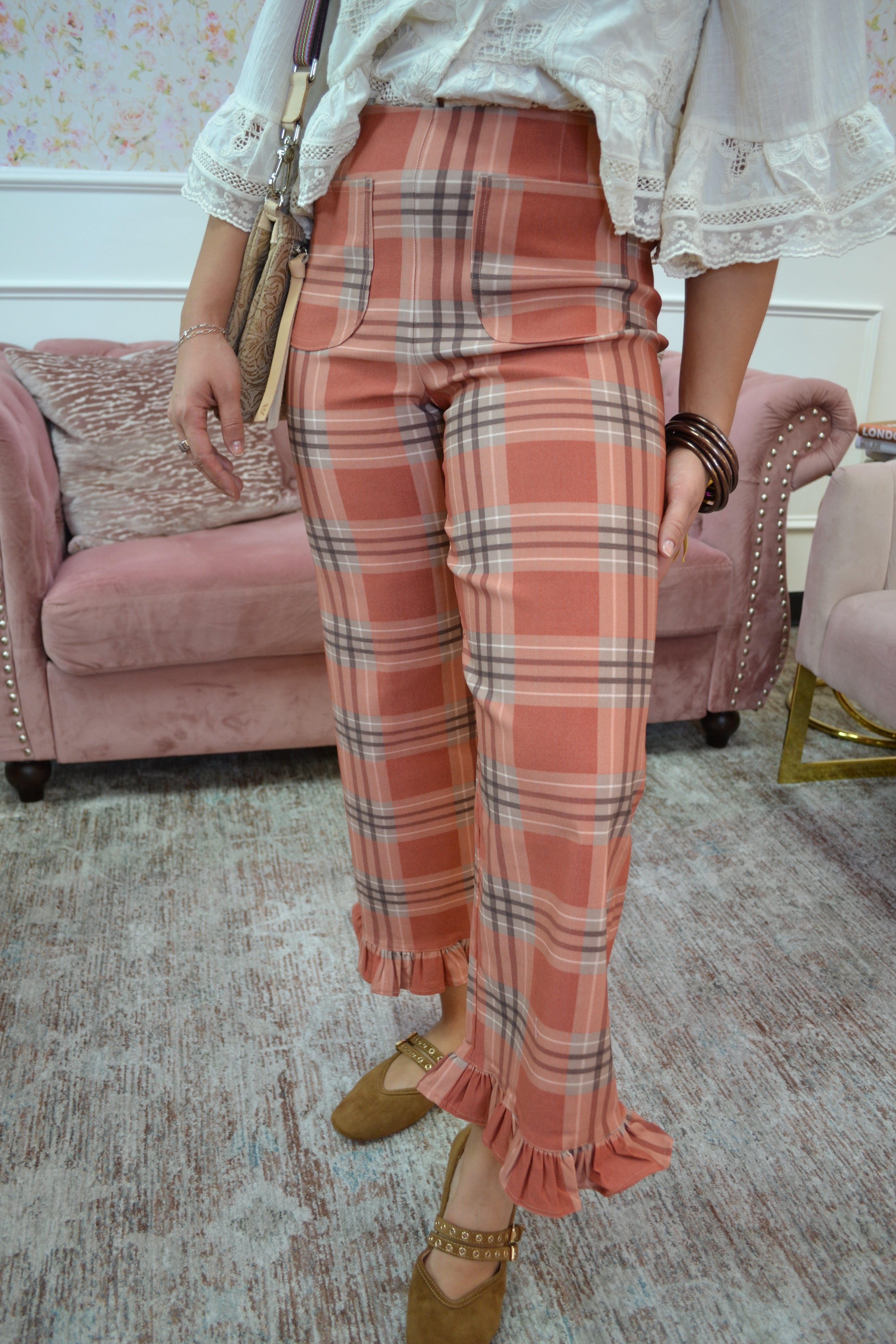 A person wearing high-waisted plaid cropped pants in a dusty pink, orange, and gray tartan pattern, paired with a white lace-trim blouse and brown shoes, standing in a living room setting.