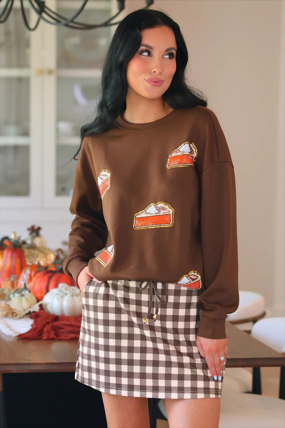 Model wearing a brown sweatshirt with embroidered pie slice patches, paired with a brown and white checkered skort, perfect for casual fall wear.