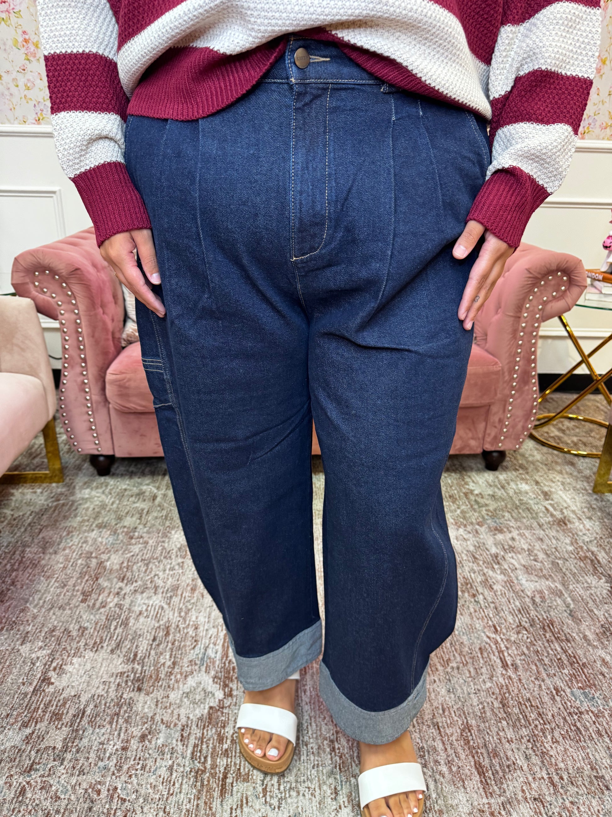 A person wearing dark blue wide-leg denim jeans with rolled cuffs, paired with a maroon and white striped sweater and white slide sandals, standing in an indoor room with pink armchairs.