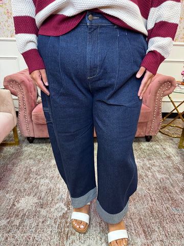 A person wearing dark blue wide-leg denim jeans with rolled cuffs, paired with a maroon and white striped sweater and white slide sandals, standing in an indoor room with pink armchairs.