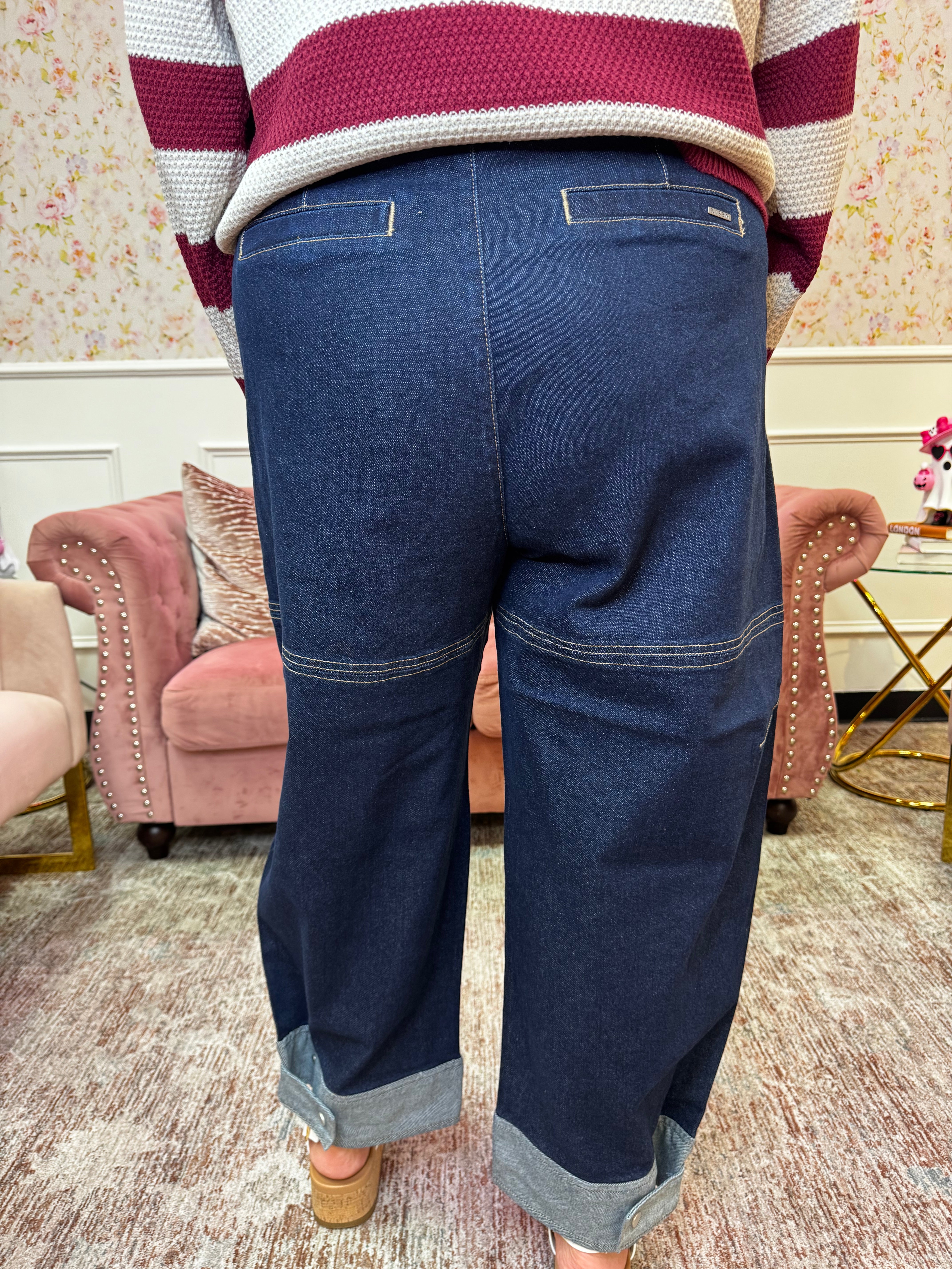 A person shown from the back wearing dark blue wide-leg denim jeans with cuffed hems and contrast stitching, paired with a striped sweater in a decorated room with pink armchairs.
