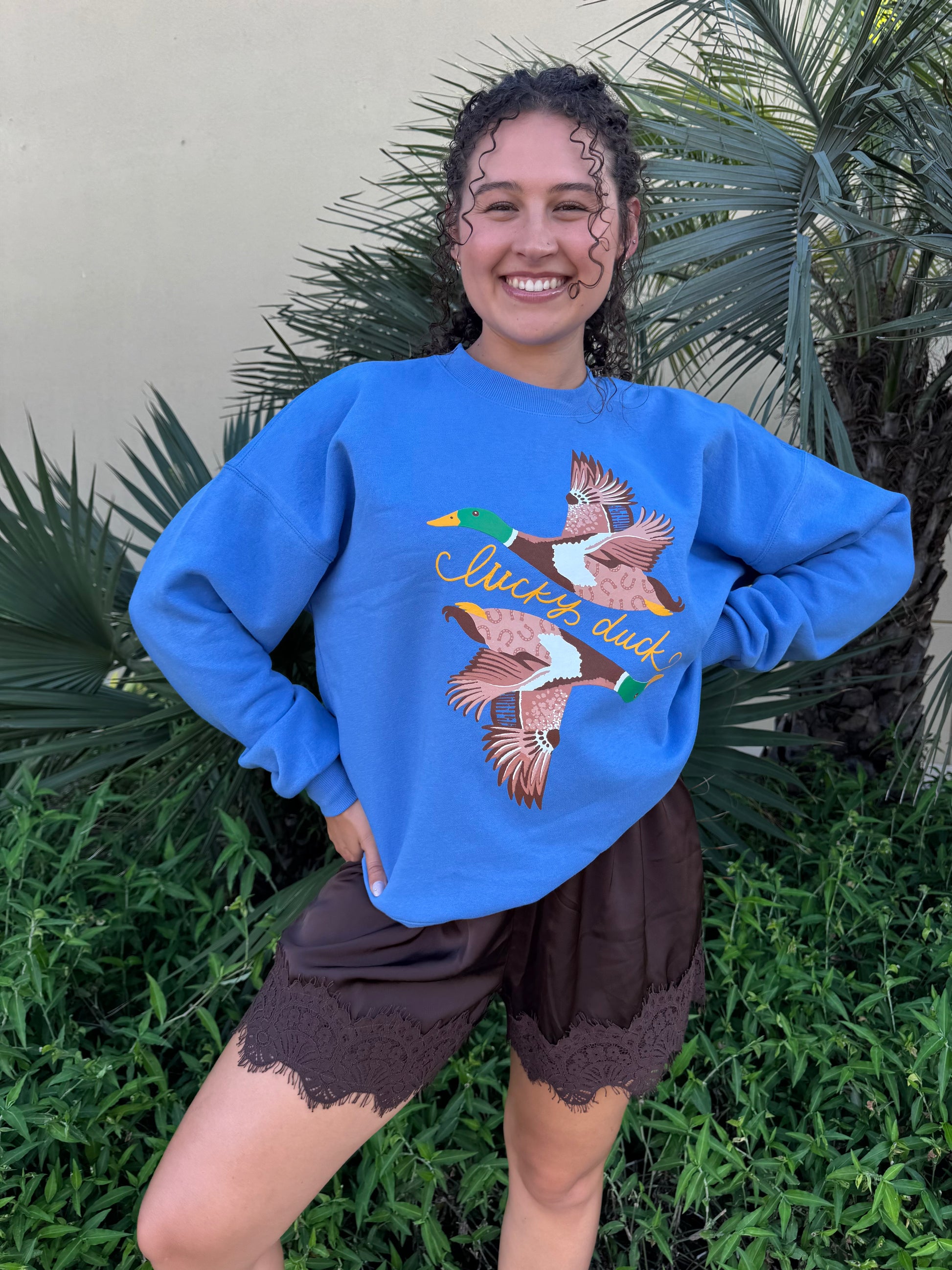 Young woman smiling outdoors wearing a bright blue duck graphic sweatshirt with a relaxed fit, paired with brown lace-trim shorts, surrounded by lush green plants.