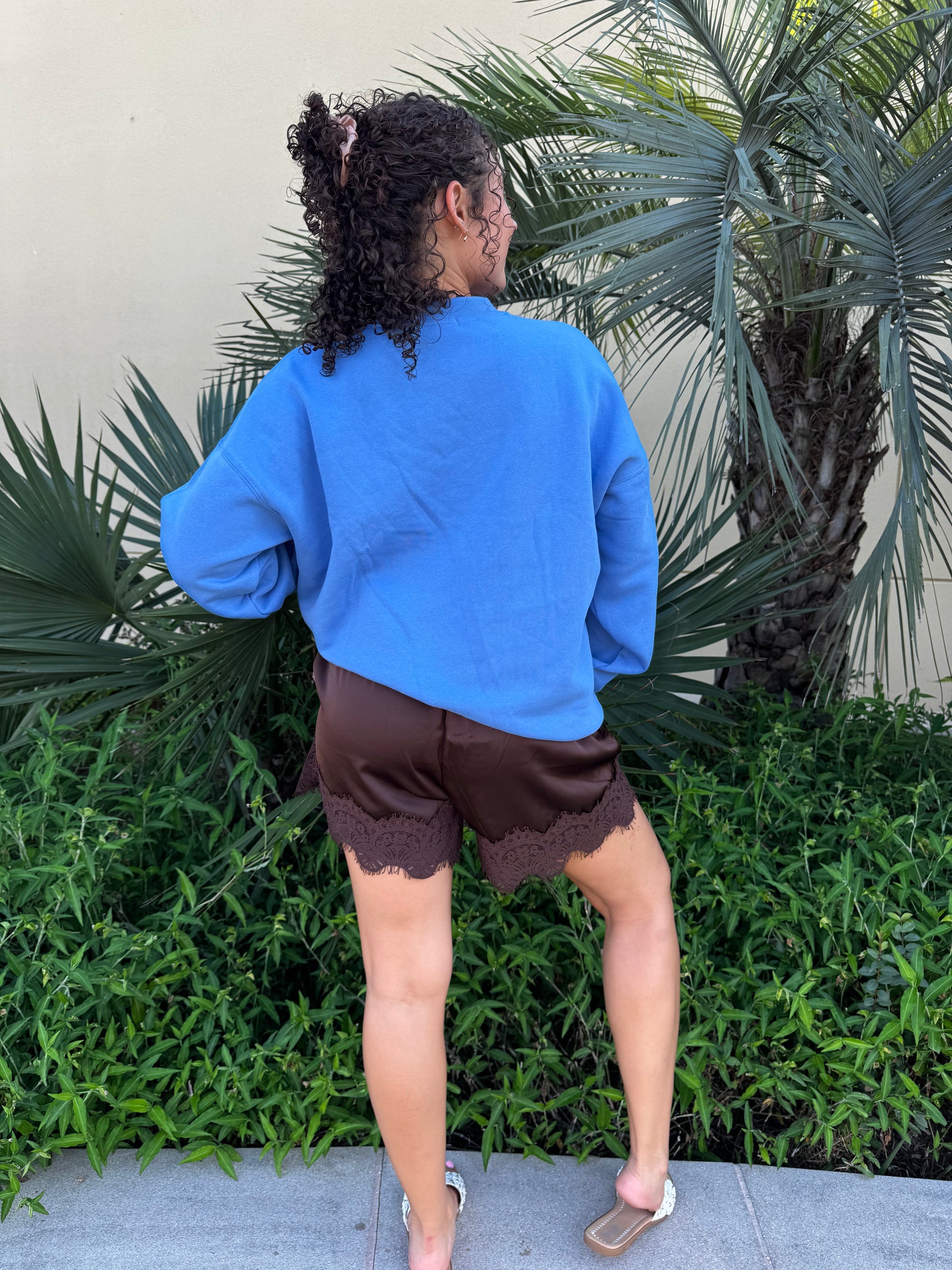 Woman with curly hair in blue duck graphic sweatshirt, standing near palm trees, smiling and looking over her shoulder.