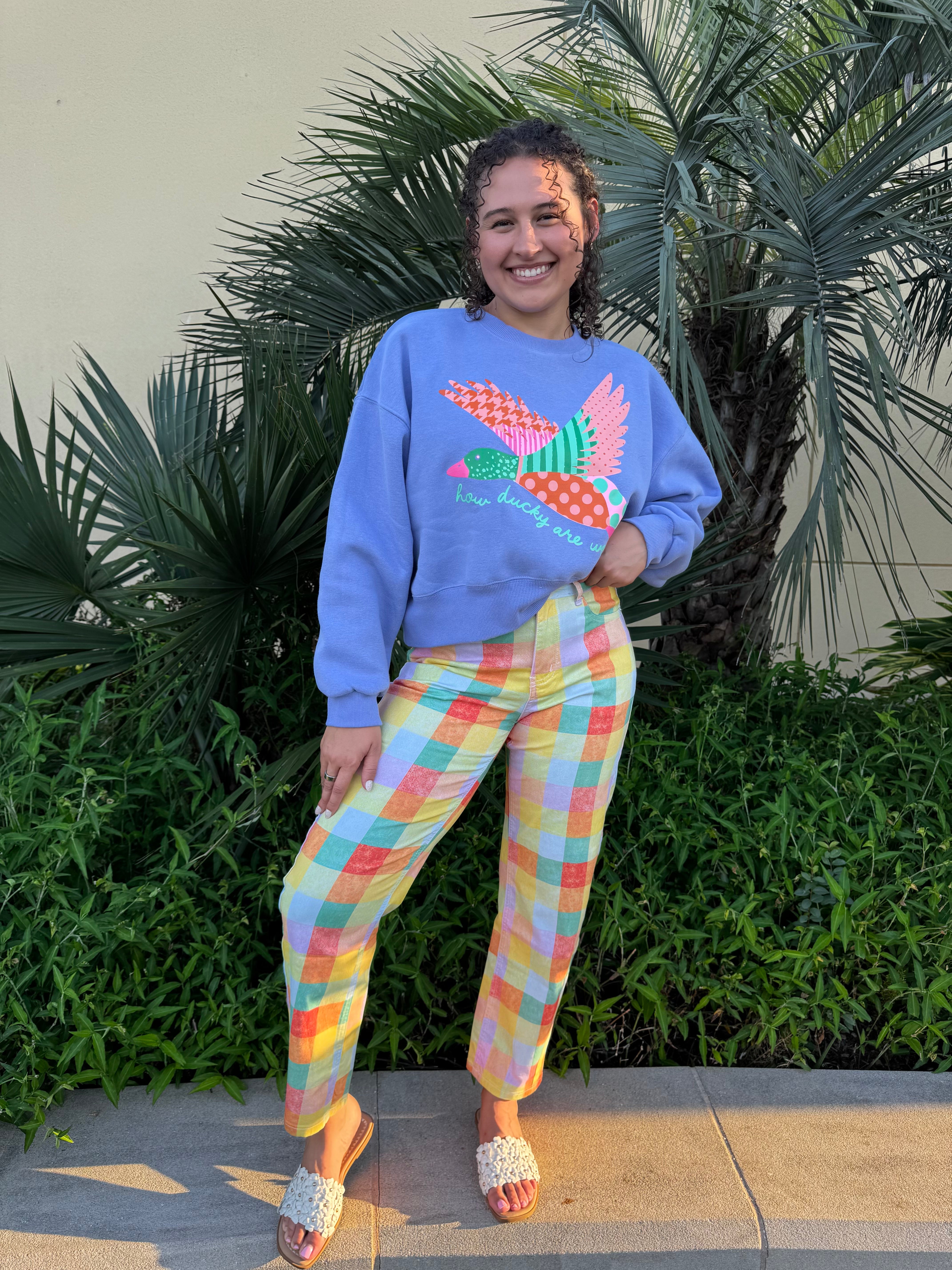 Smiling woman wearing a blue cropped sweatshirt with colorful duck graphic and pastel plaid pants, standing outdoors near palm plants.