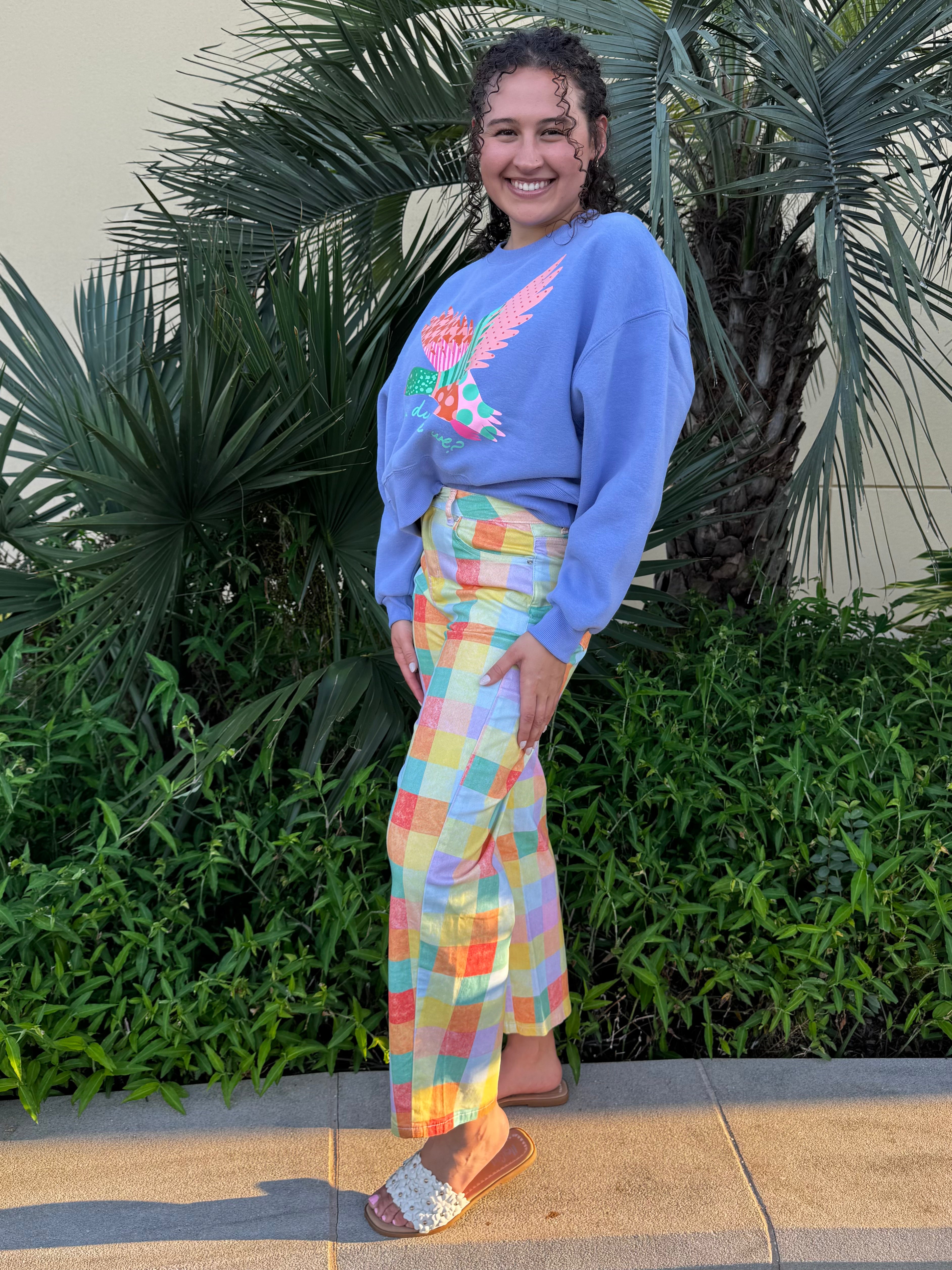 Close-up of woman in blue sweatshirt with duck graphic and pastel plaid pants, smiling with palm plants in background.