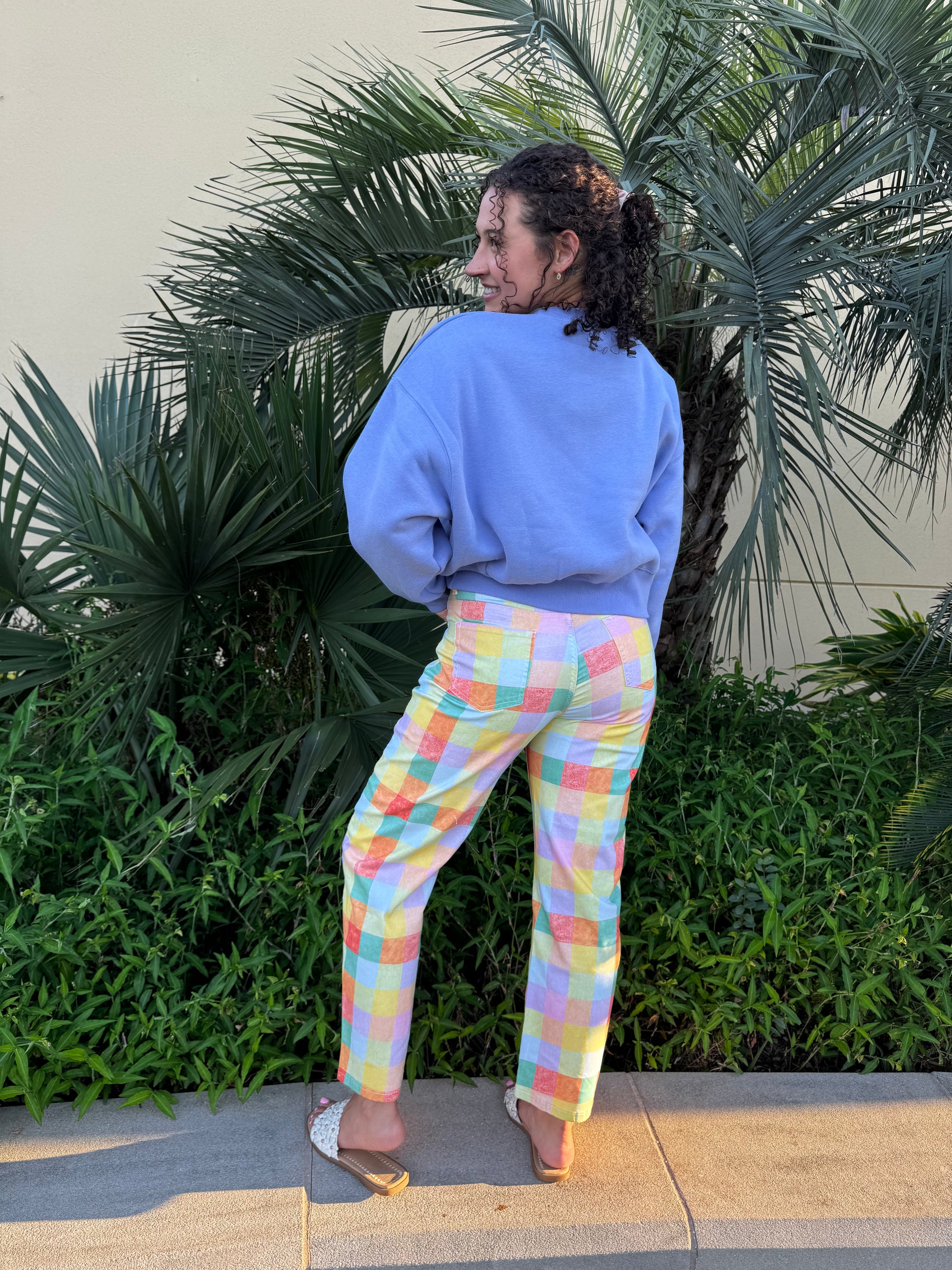 Back view of woman wearing pastel plaid high-rise barrel pants and blue sweatshirt, standing near tropical plants outdoors.