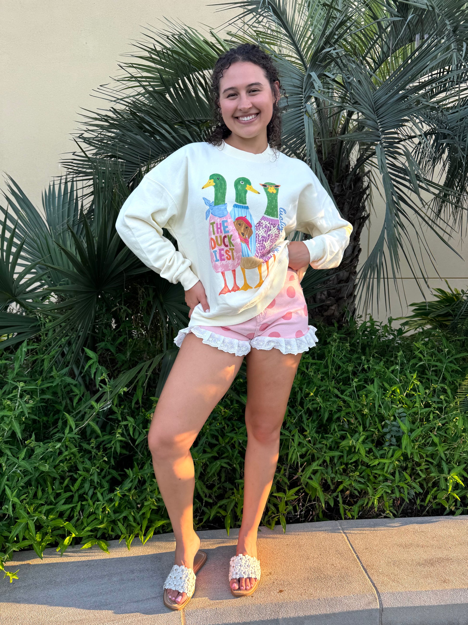 Young woman smiling outdoors wearing high-rise pink polka dot shorts with white lace trim, cream ruffle denim shorts, and a white sweatshirt, standing near lush green plants.