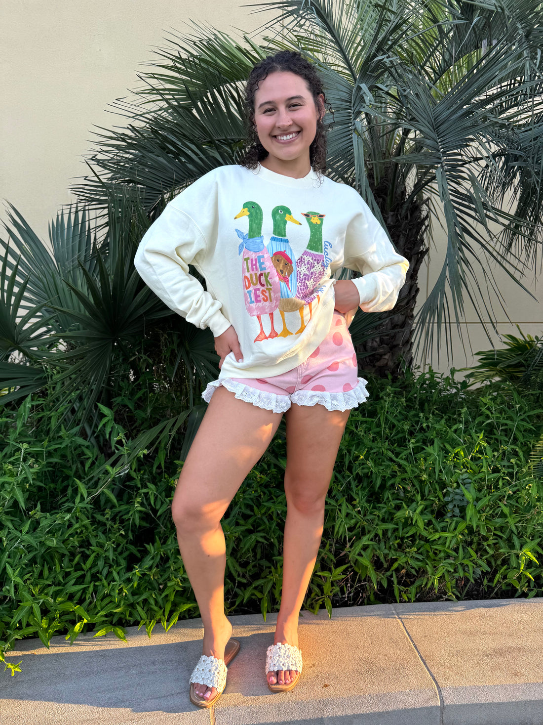 Young woman smiling outdoors wearing a cream crewneck sweatshirt with colorful duck graphic and pink polka dot shorts, standing near lush green plants.