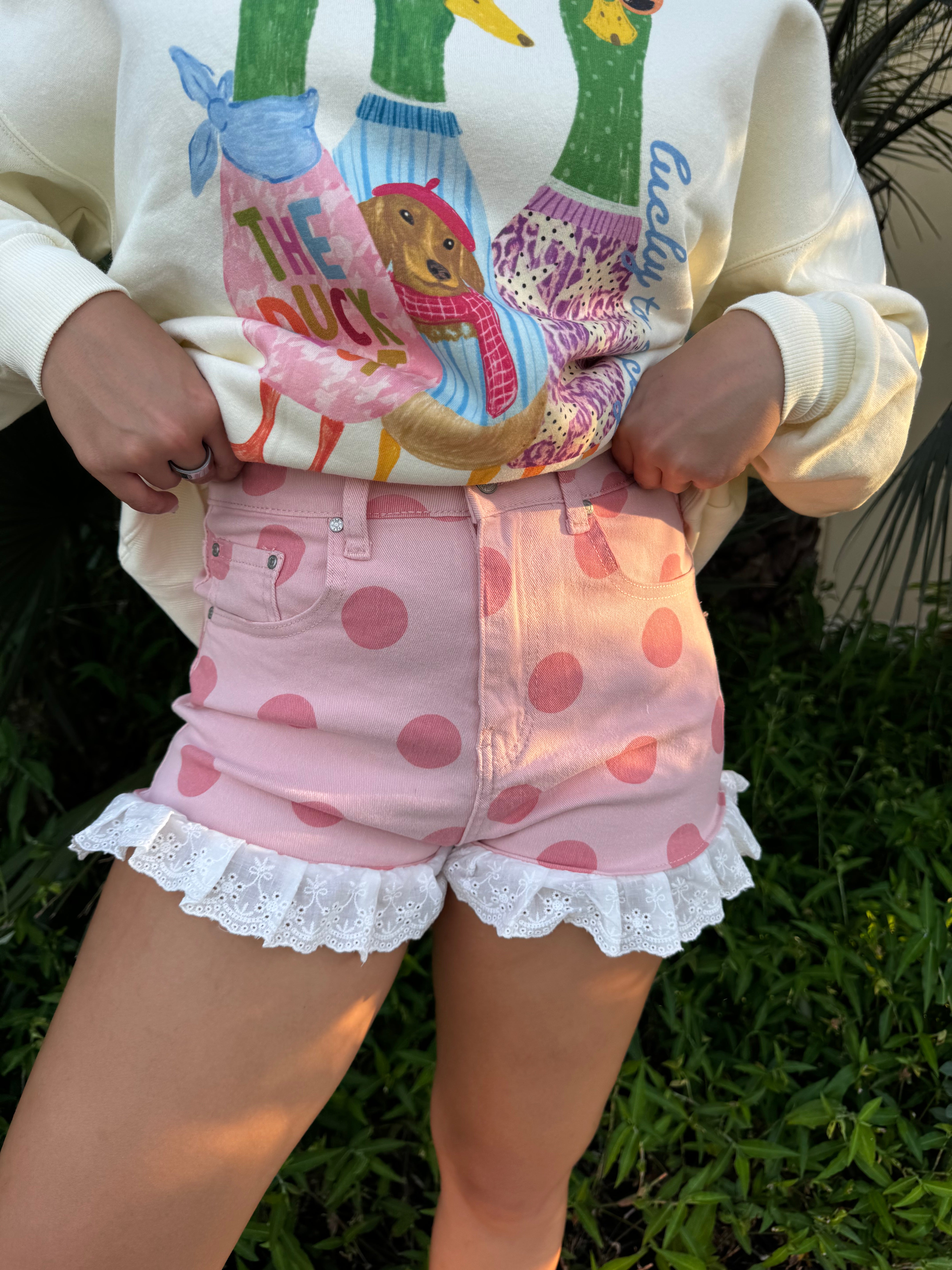 Close-up of cream ruffle denim shorts with pink polka dots and white lace trim, worn by a woman with hands in pockets, standing among green plants.