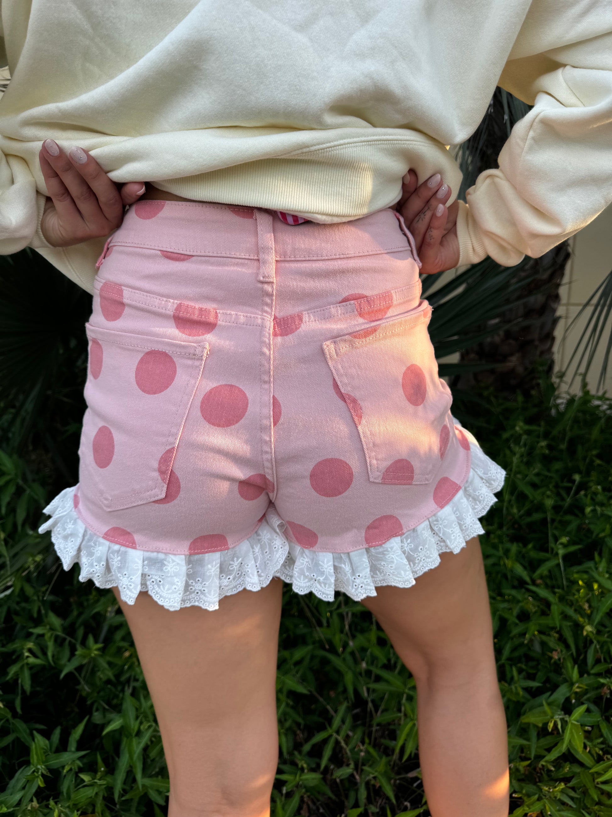 Woman in cream ruffle denim shorts with pink polka dots and white lace trim, smiling and posing outdoors near tropical plants, wearing a cream sweatshirt.