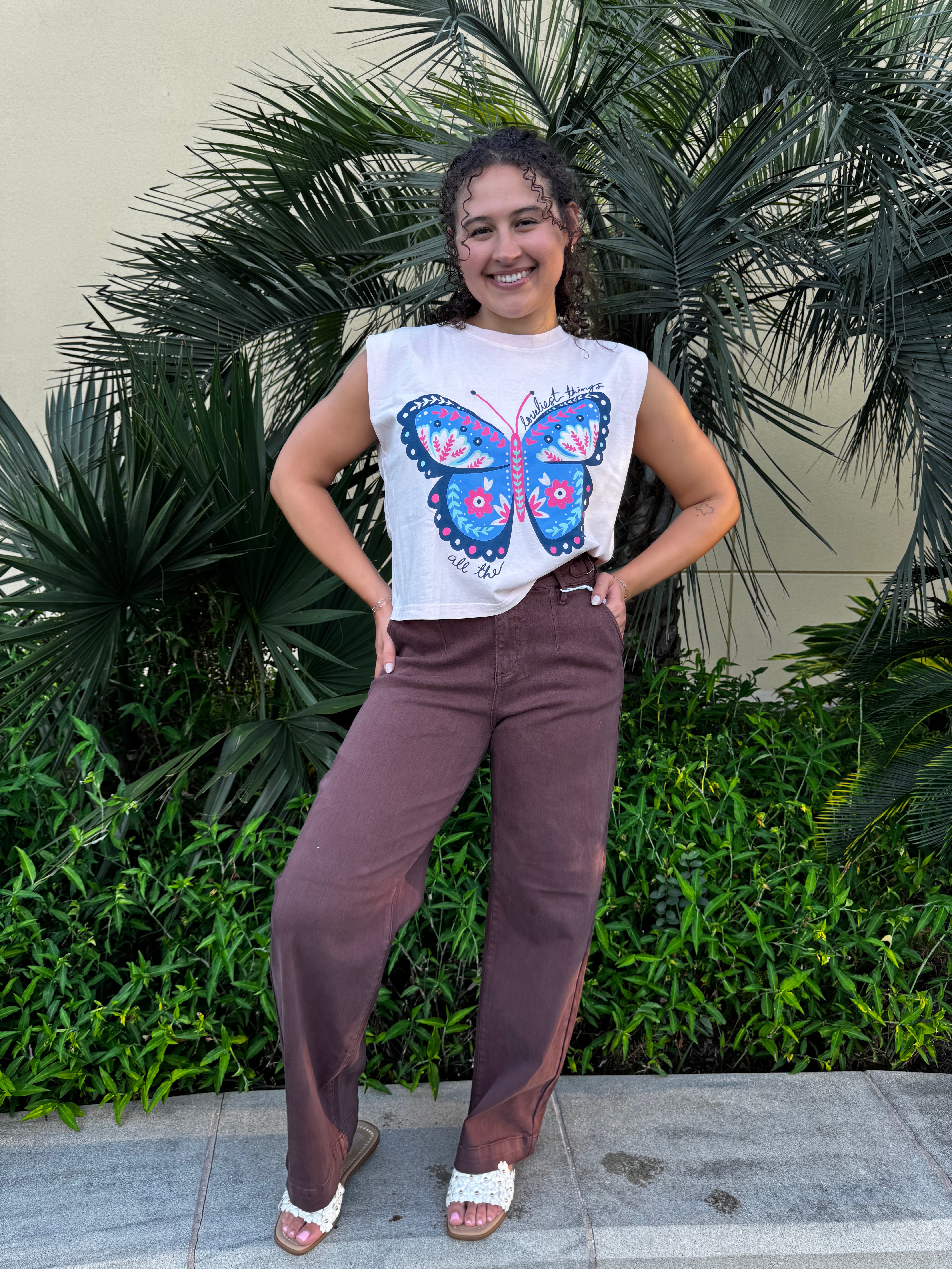 Young woman smiling in front of lush green tropical plants, wearing a white sleeveless graphic tank top with a colorful butterfly design and brown wide-leg pants