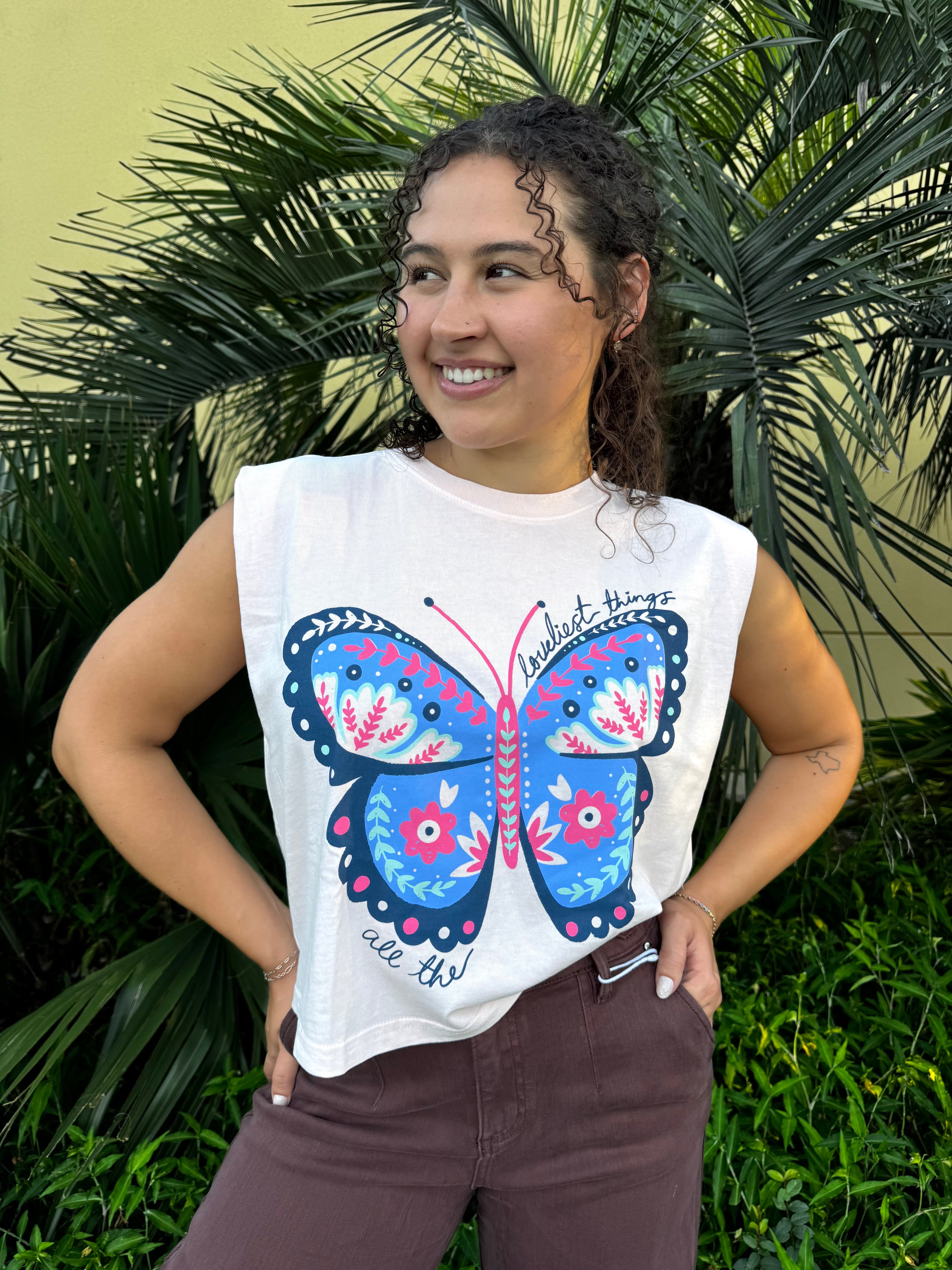 Young woman with curly hair wearing a white butterfly graphic tank top with pink, blue, and floral details, standing outdoors with green foliage background