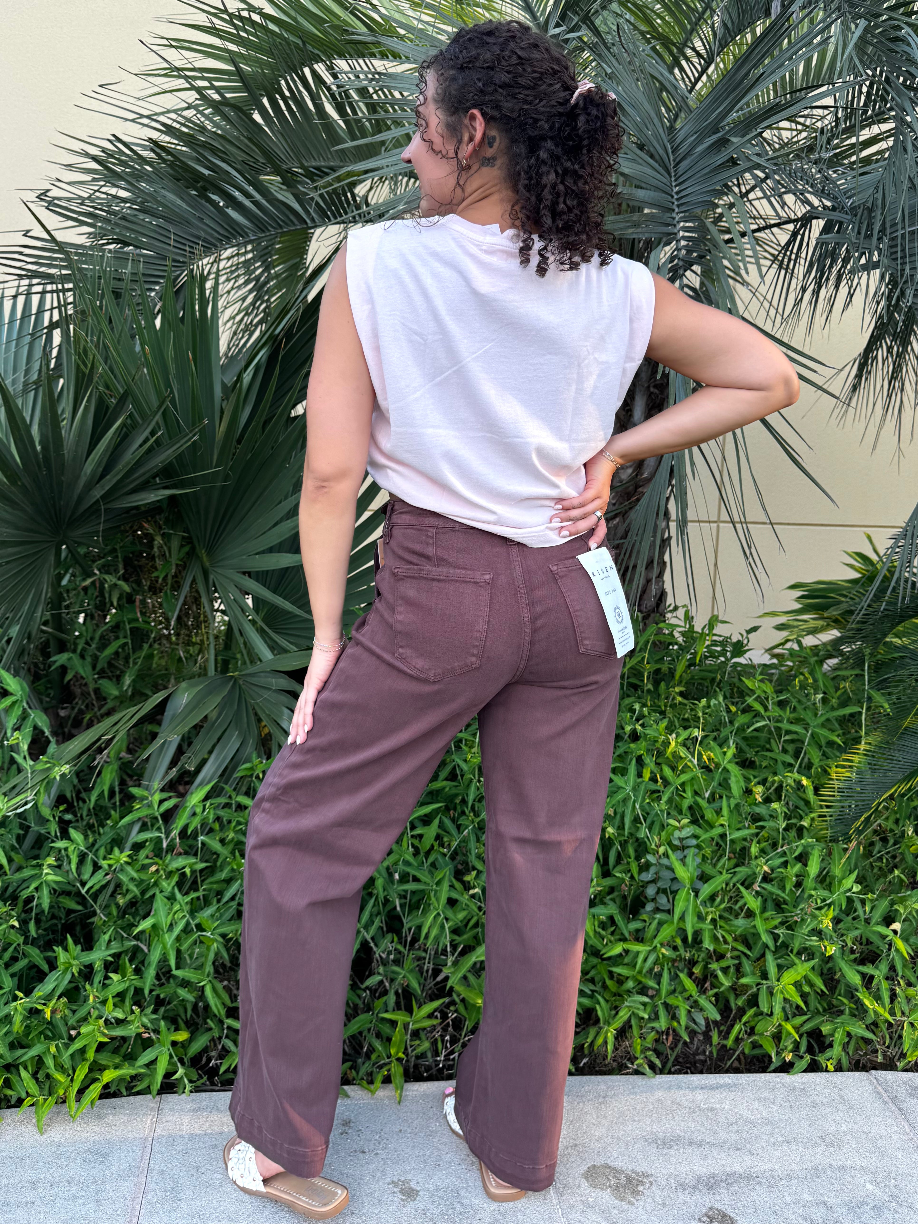 Woman with curly hair wearing a sleeveless white cotton tank top and high-waisted brown pants, standing outdoors with lush green tropical plants in the background.