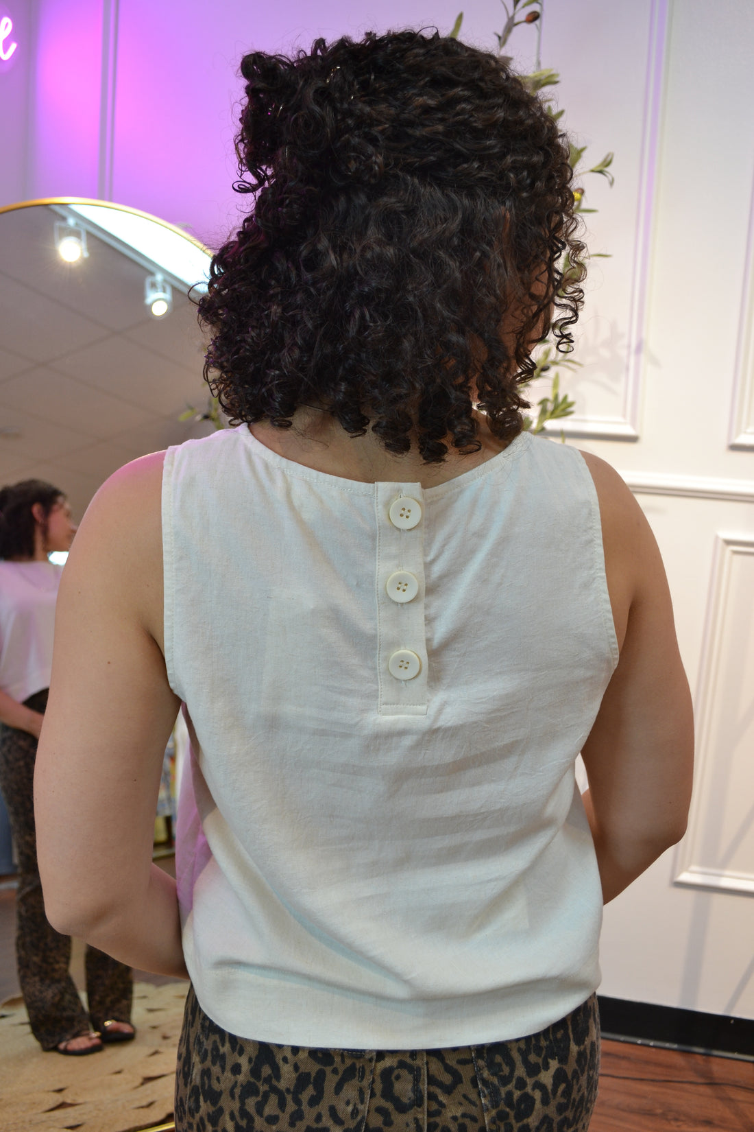 Woman wearing a white sleeveless top and leopard print pants standing in a room with a mirror and plant.