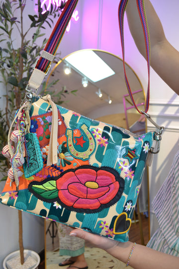 Colorful embroidered floral crossbody handbag with a rainbow strap, horseshoe and star patches, glossy turquoise geometric print, tassel charm, and boho-inspired design displayed in a boutique.