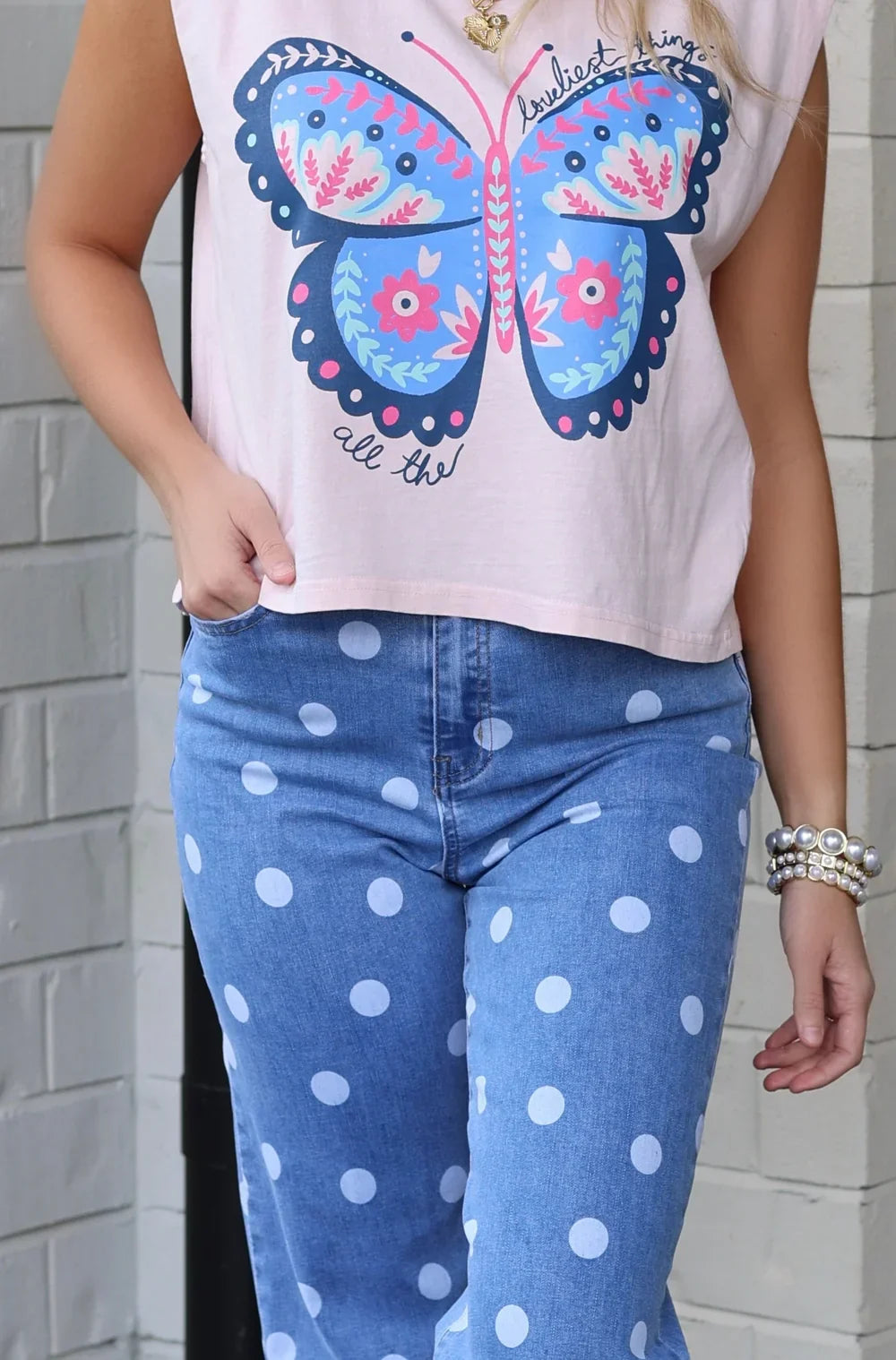 Women's light pink graphic tank top featuring a colorful butterfly design with floral details and handwritten text, paired with polka dot jeans