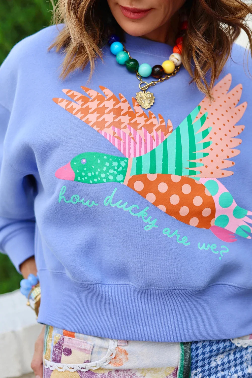 Woman in blue sweatshirt with duck graphic and pastel plaid pants, showing back view with palm plants behind her.