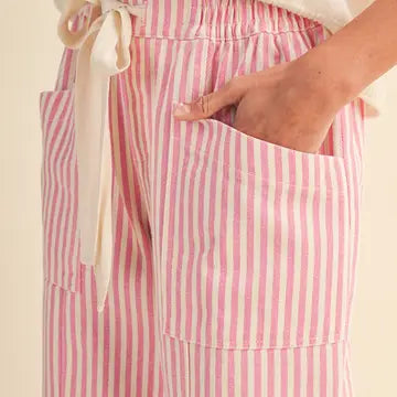 Pink and white vertically striped linen pants with an elastic waist and cream-colored fabric tie, shown on a model with a hand in the pocket.